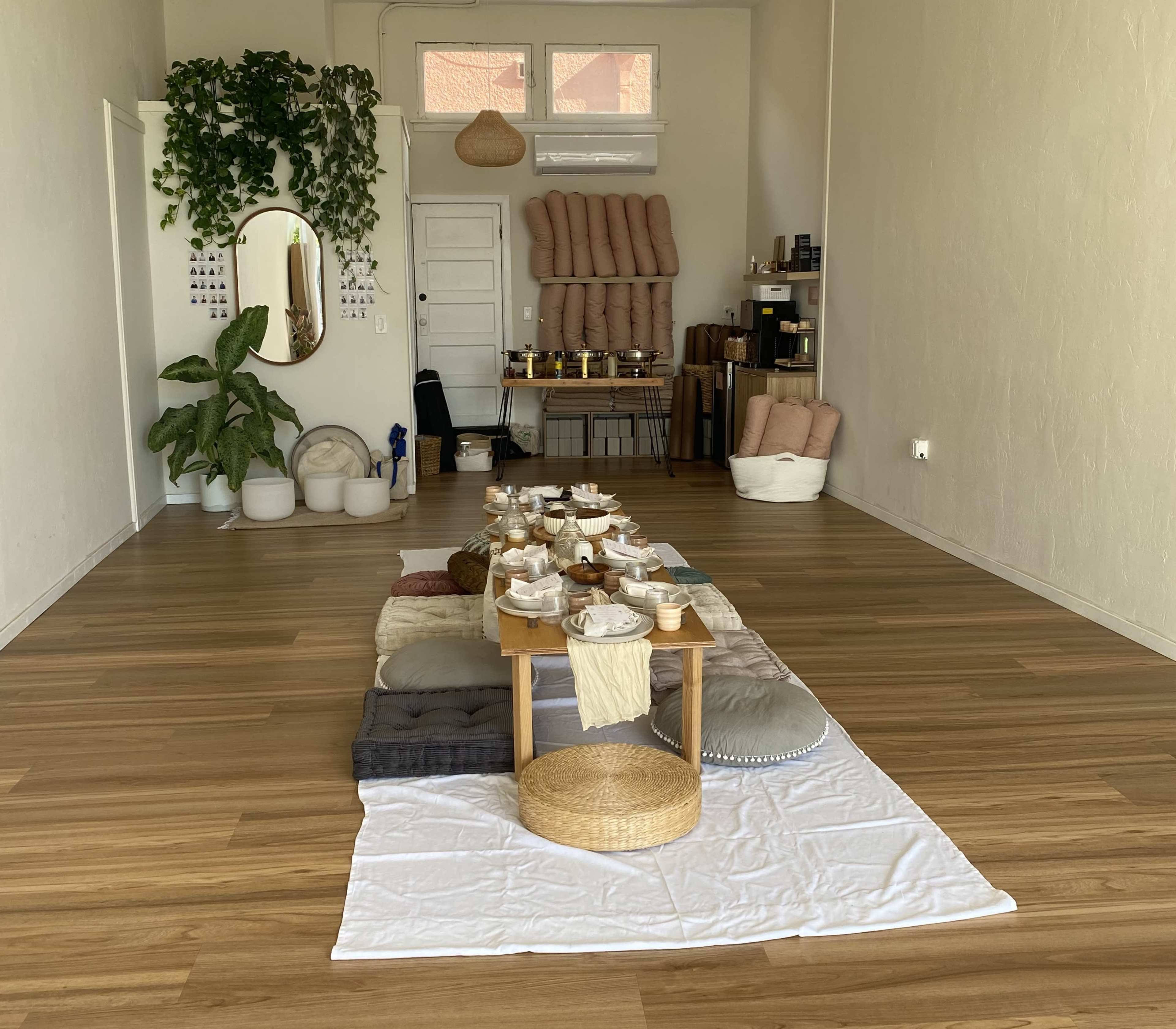 The image shows a spacious interior with wooden flooring, a long table set with dishes and decor, surrounded by plants and various furnishings against the walls.