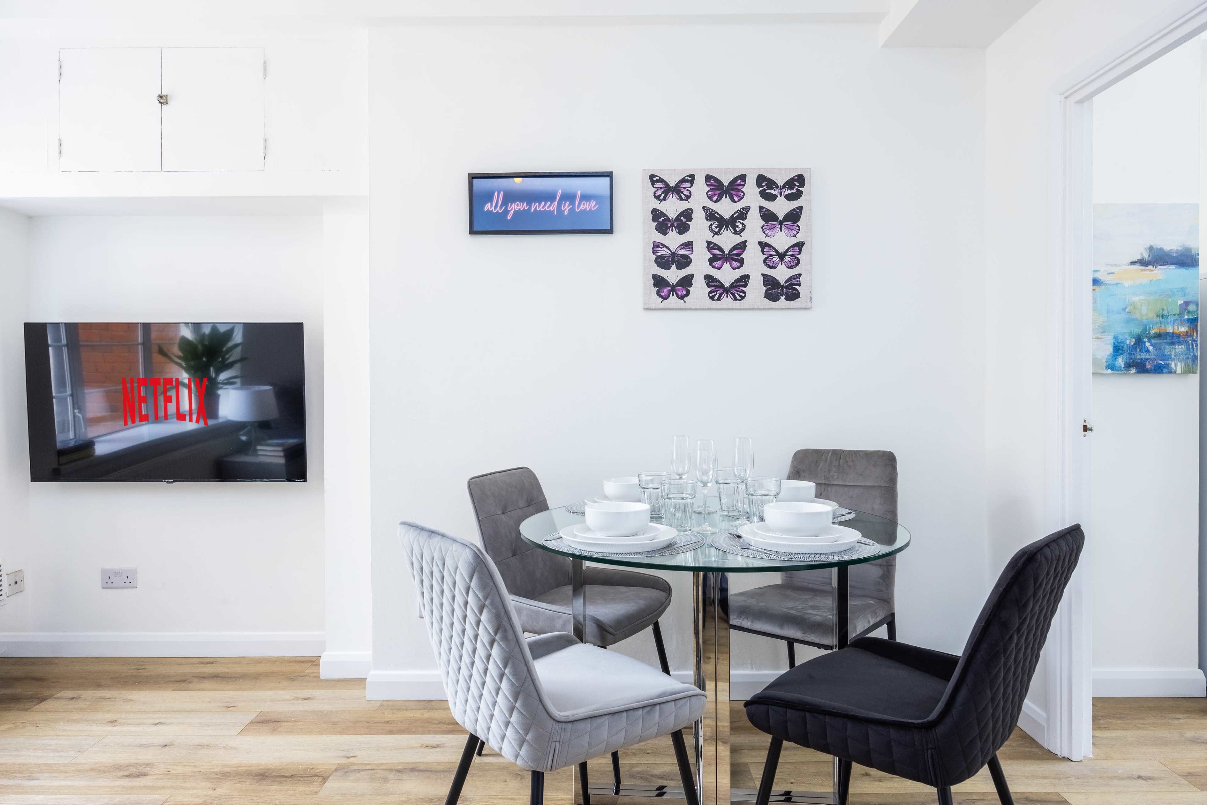 The image shows a dining area with a glass circular table set for four, surrounded by four chairs, and a wall-mounted television displaying the Netflix logo.