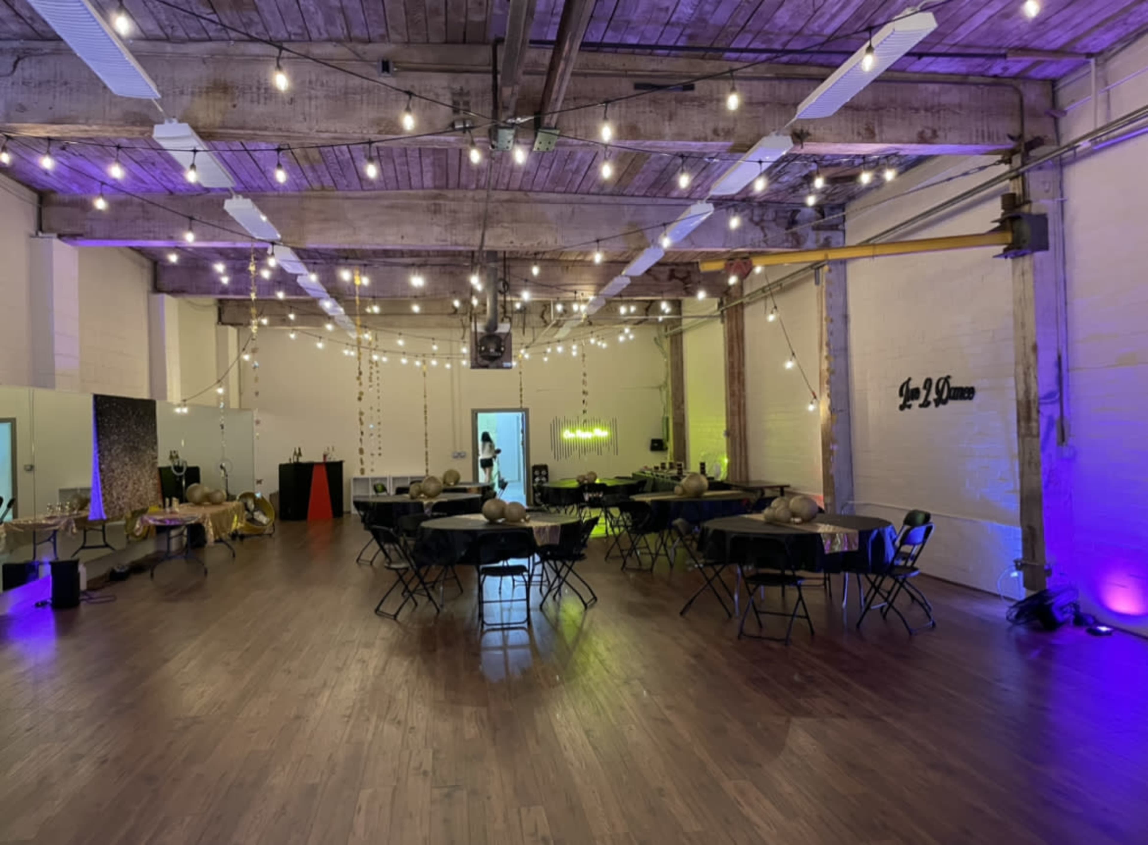 A spacious event room with tables set up, string lights overhead, and a bar area in the background.