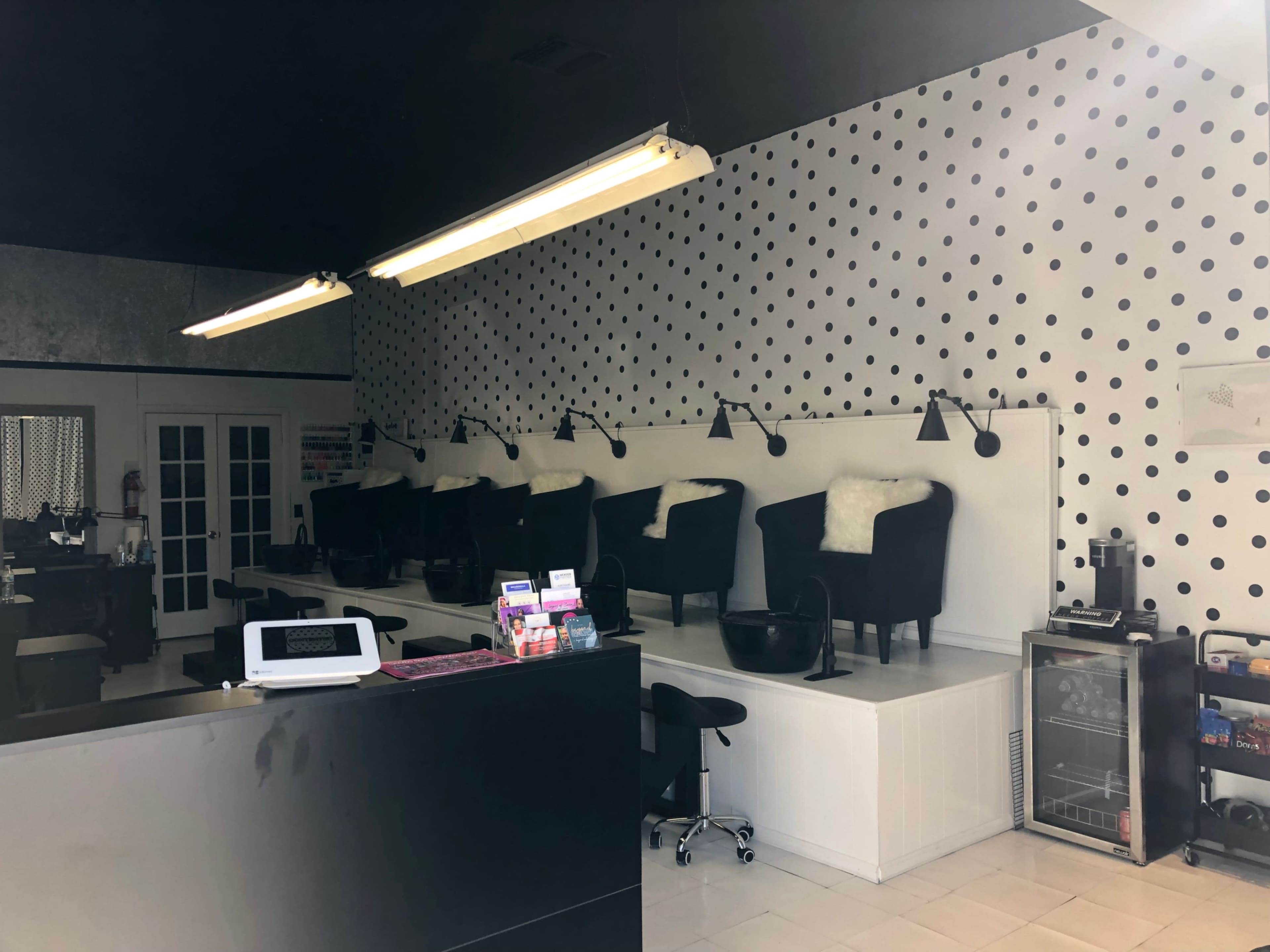 A nail salon with black chairs along a white wall decorated with black polka dots and a reception counter in the foreground.