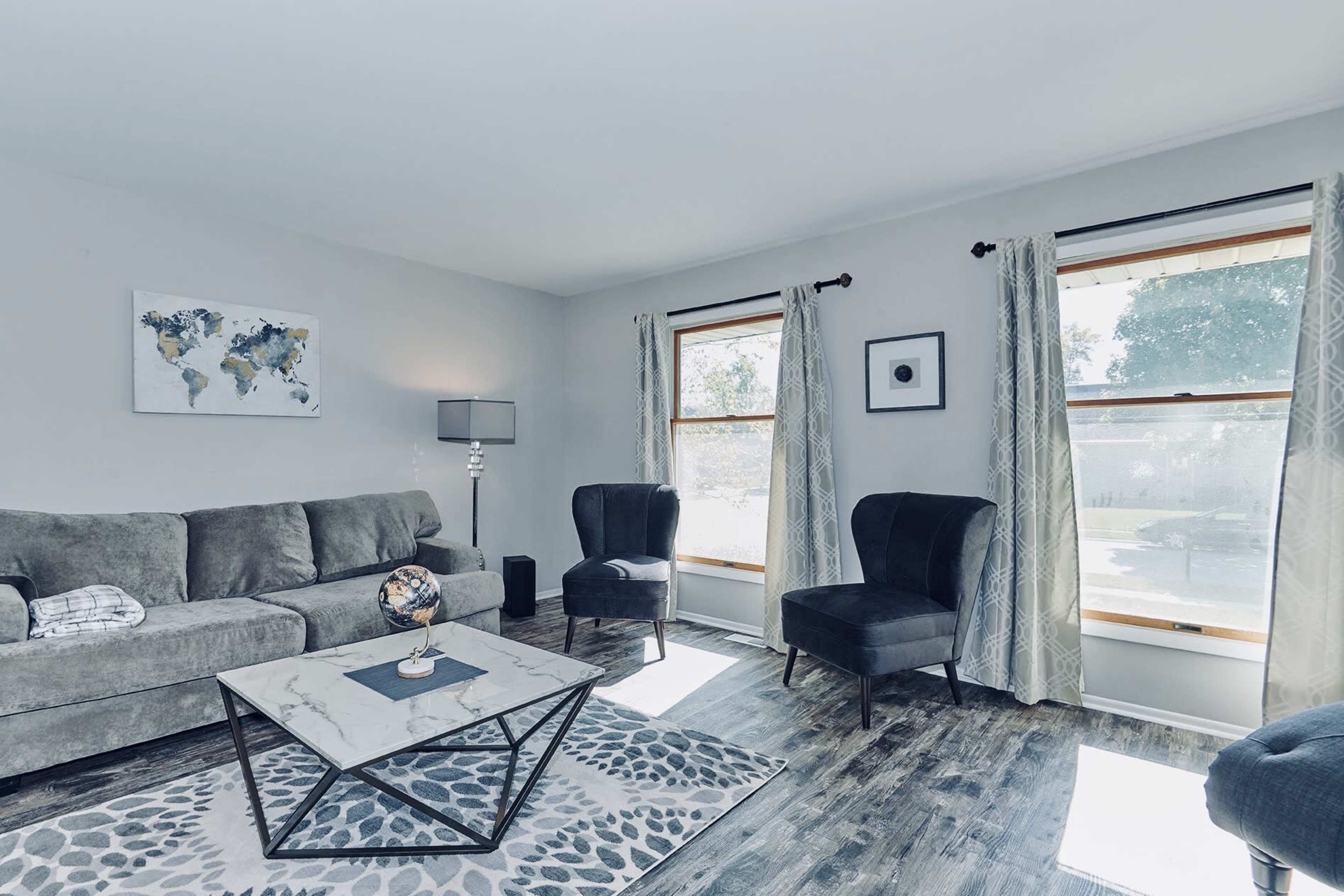 The living room features a gray sofa, two dark chairs, a marble coffee table, and large windows with light curtains.