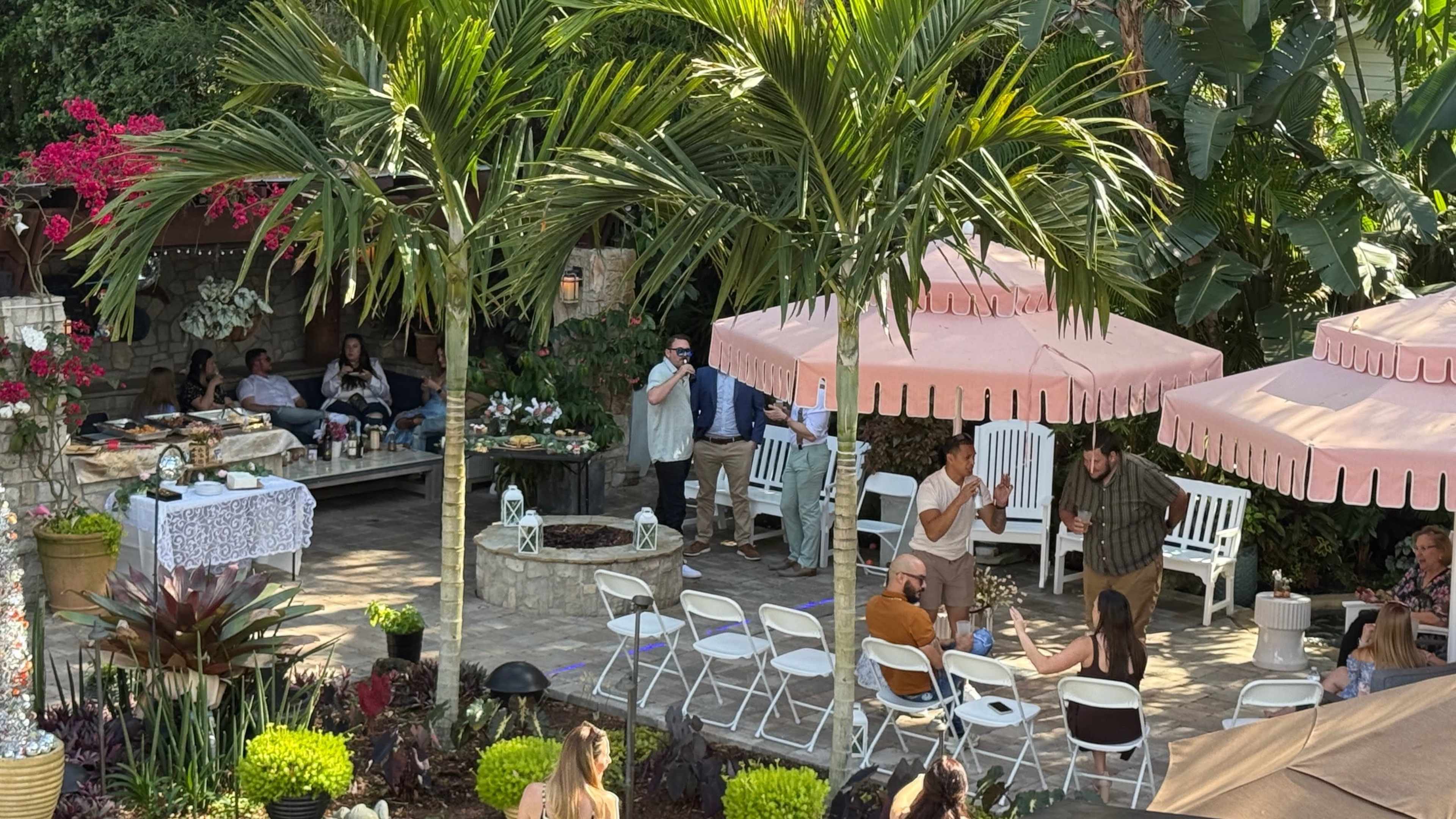 A group of people gathers in a lush garden setting with palm trees and pink umbrellas, engaging in conversation amidst tables set with food and decorations.