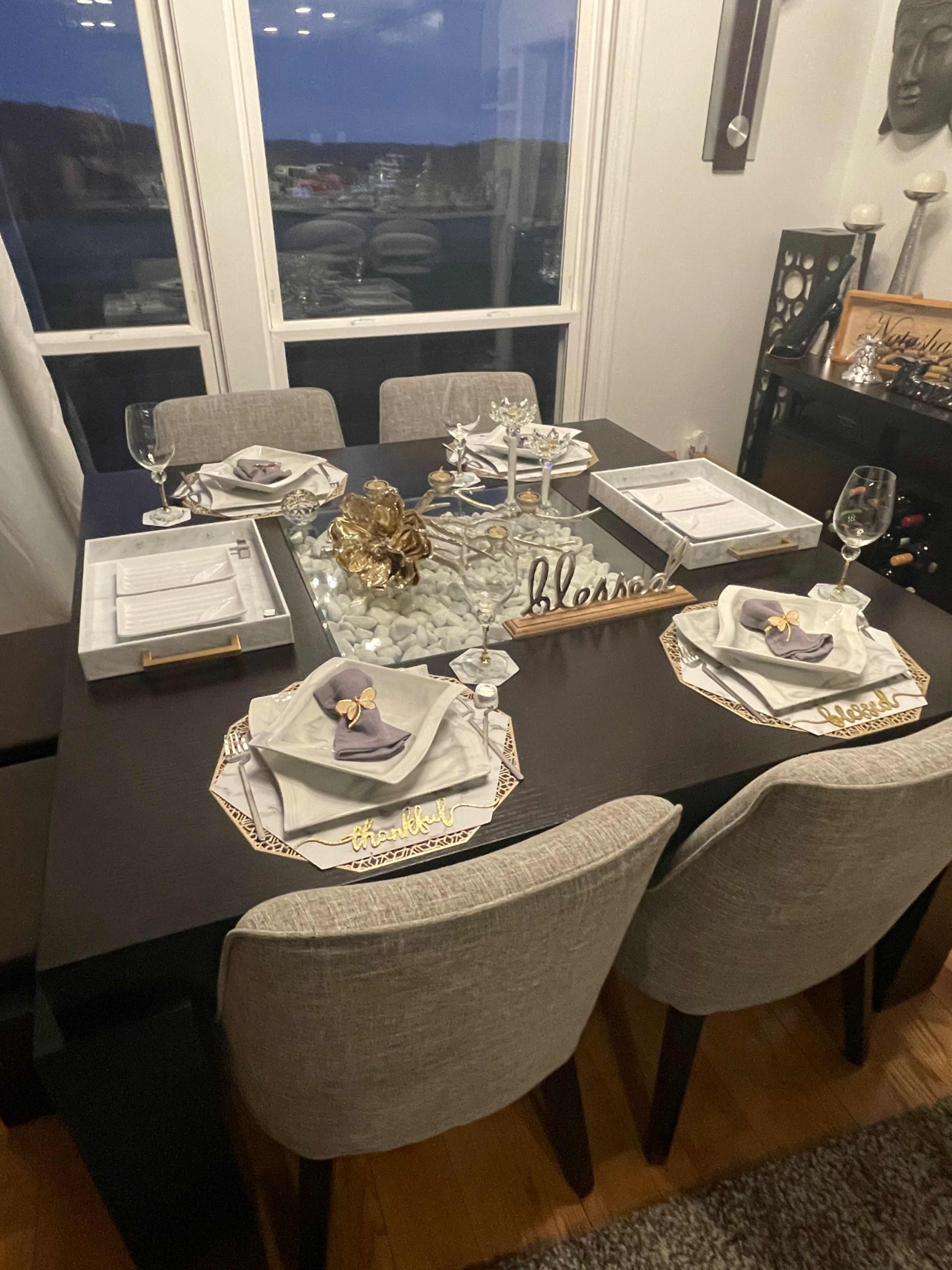 A dining table is set with elegant place settings, including folded napkins, glassware, and a decorative centerpiece, near a window displaying a nighttime view.