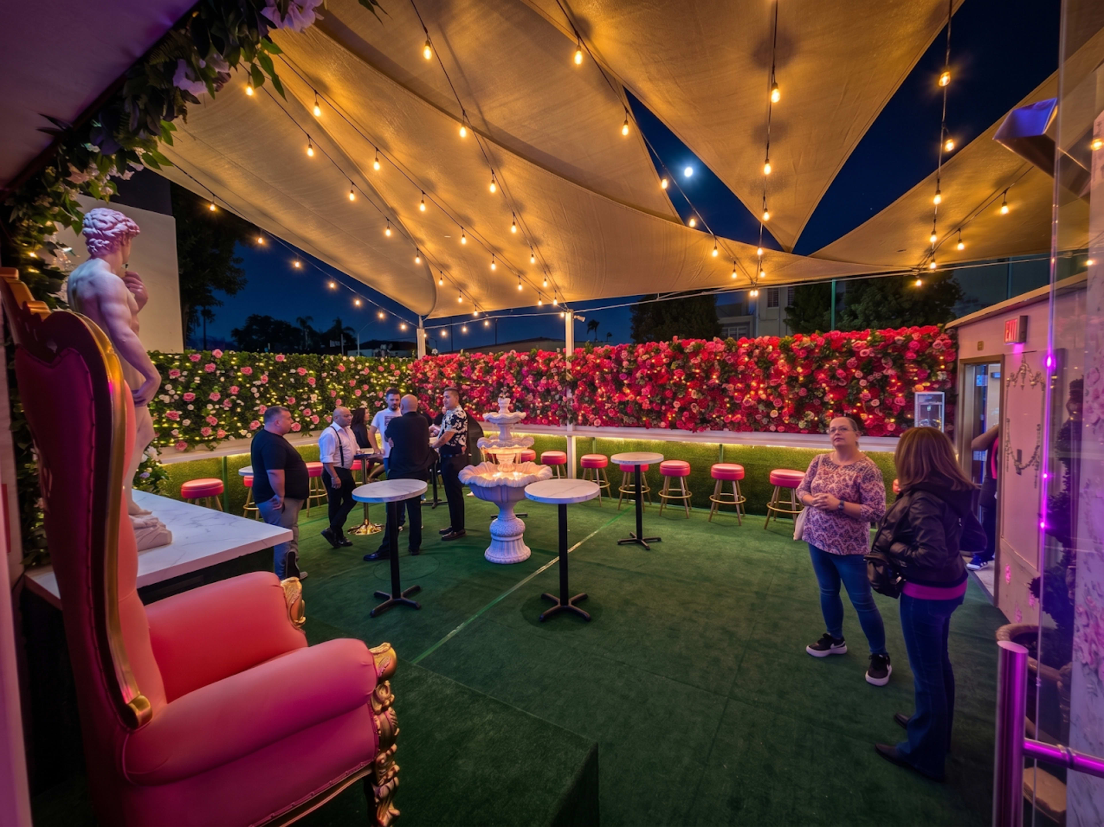 The scene shows a lively outdoor venue with a green carpet, elegant seating, and a backdrop of vibrant flowers, where people are socializing under string lights at dusk.