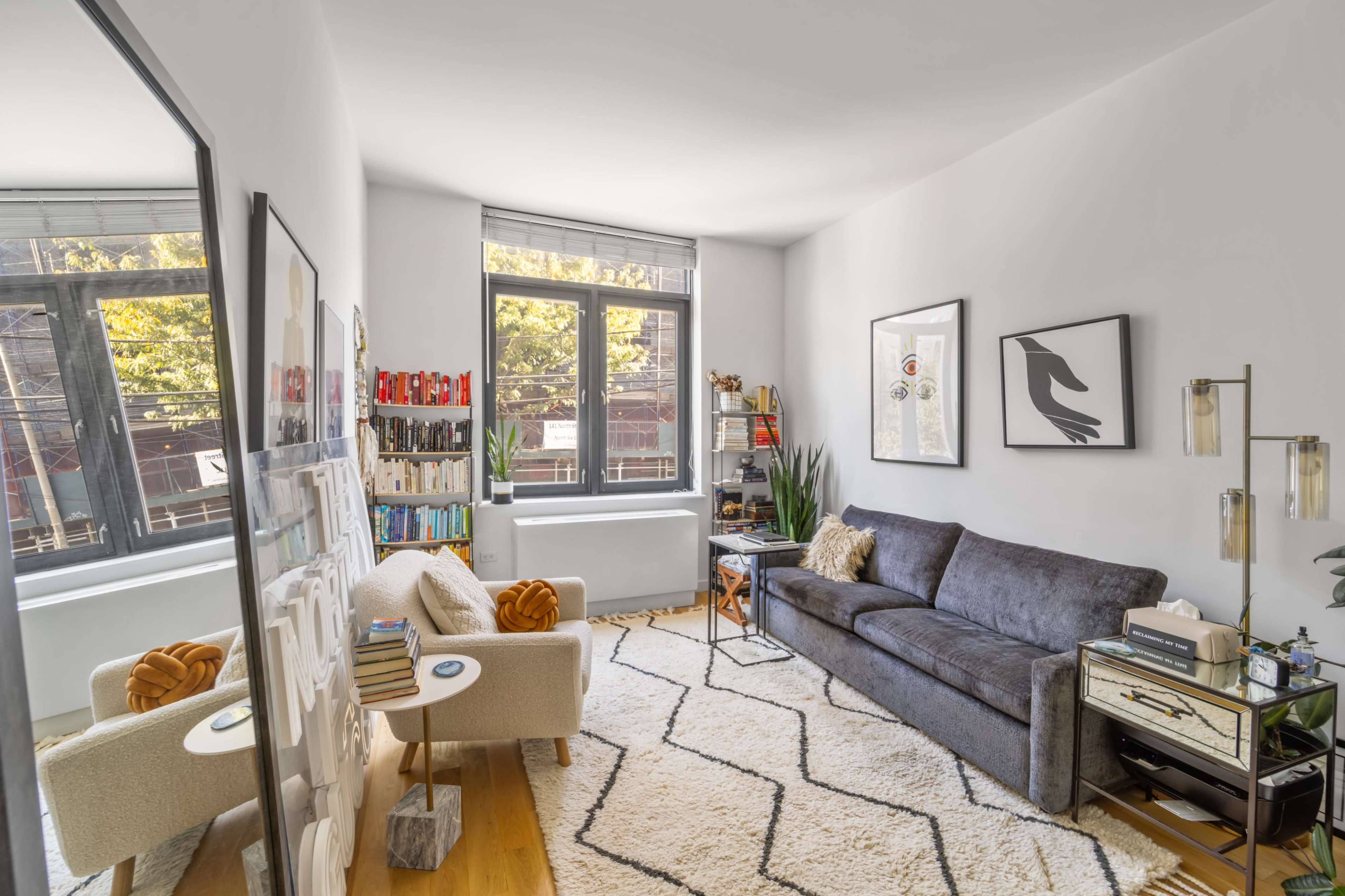 The image shows a modern living room with a gray sofa, a small coffee table, bookshelves filled with books, and large windows letting in natural light.