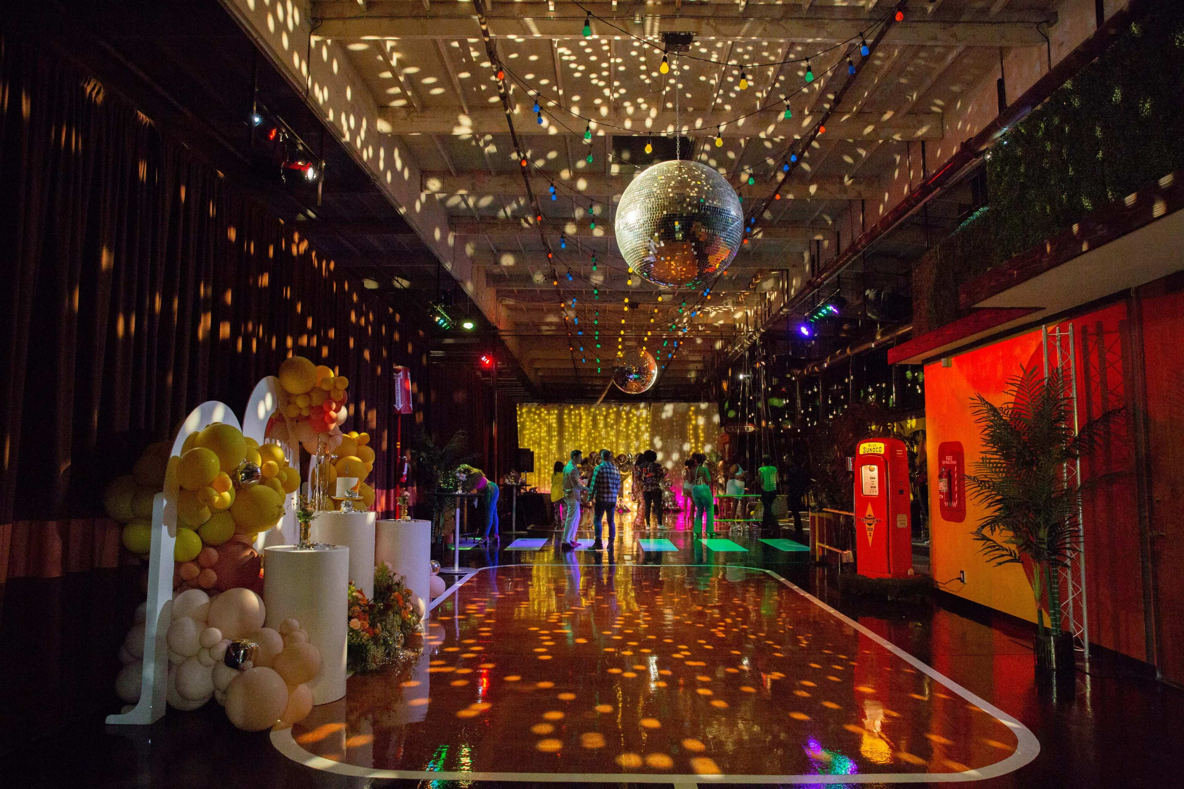 The image shows a vibrant party scene with colorful lights reflecting off disco balls and decorations, creating a festive atmosphere in a spacious venue.