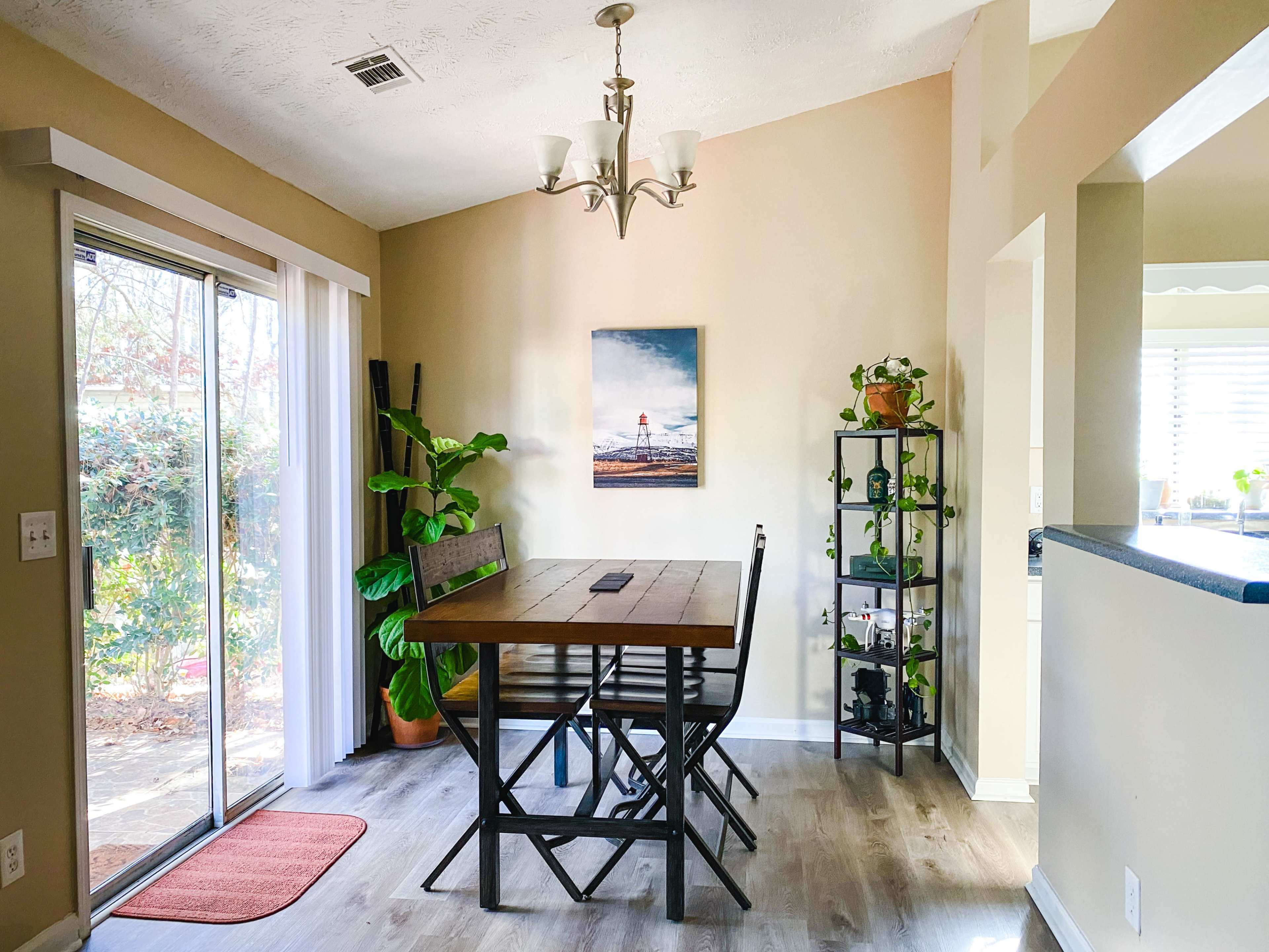 A dining area features a wooden table with four chairs, a large window with sliding glass doors, and a plant stand beside a wall-mounted art piece.