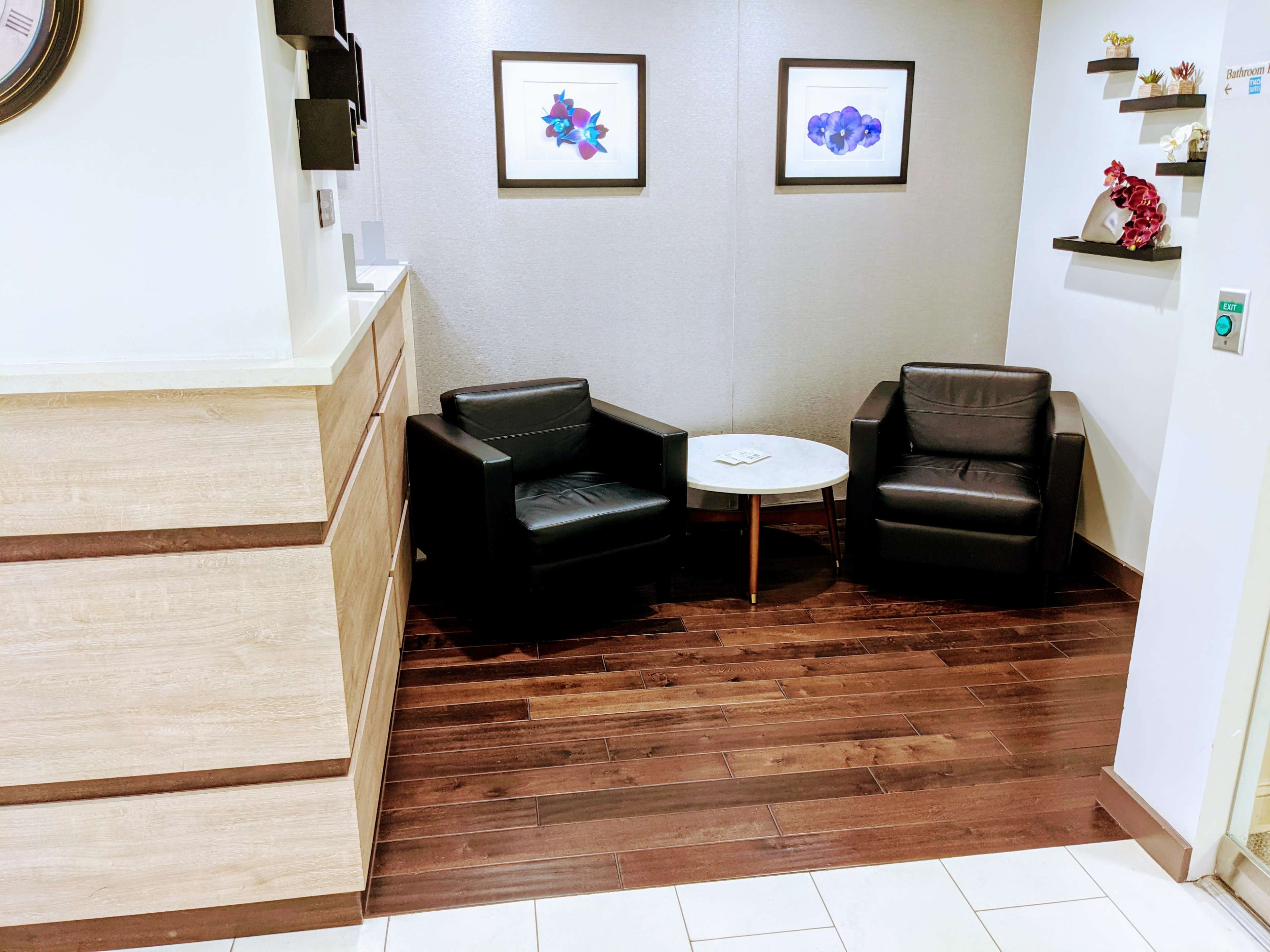 The image shows a small waiting area with two black chairs, a round table, and framed artwork on the wall.