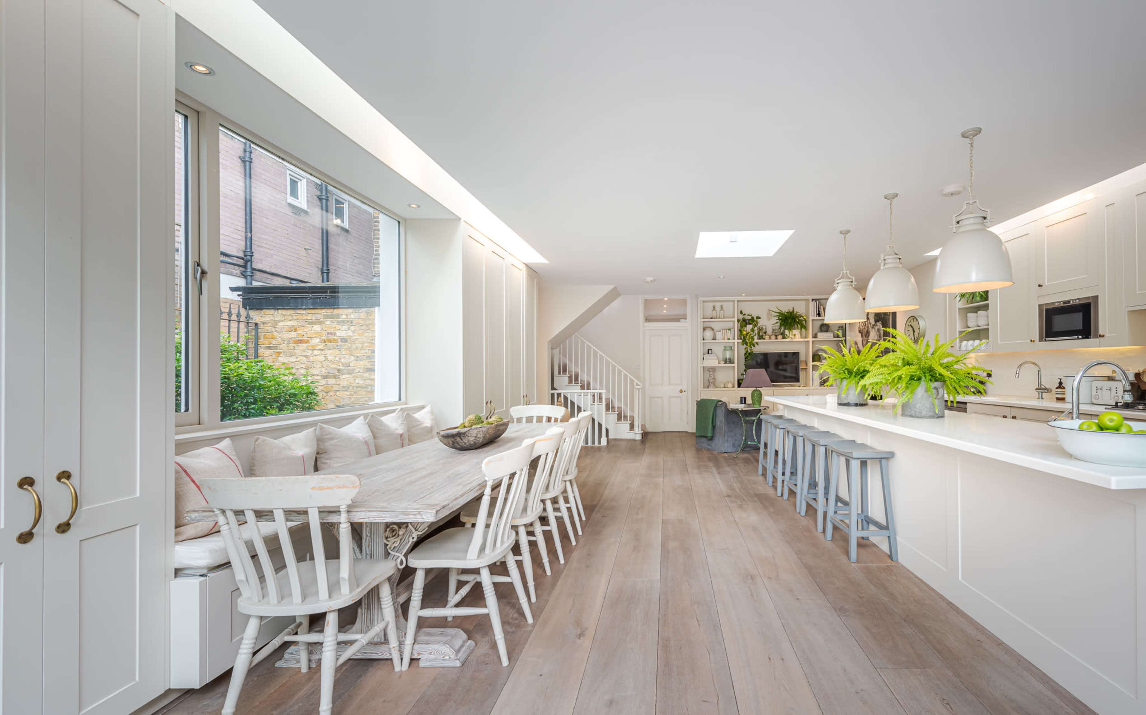 The image shows a modern kitchen and dining area with a long table, white cabinetry, and large windows allowing natural light.