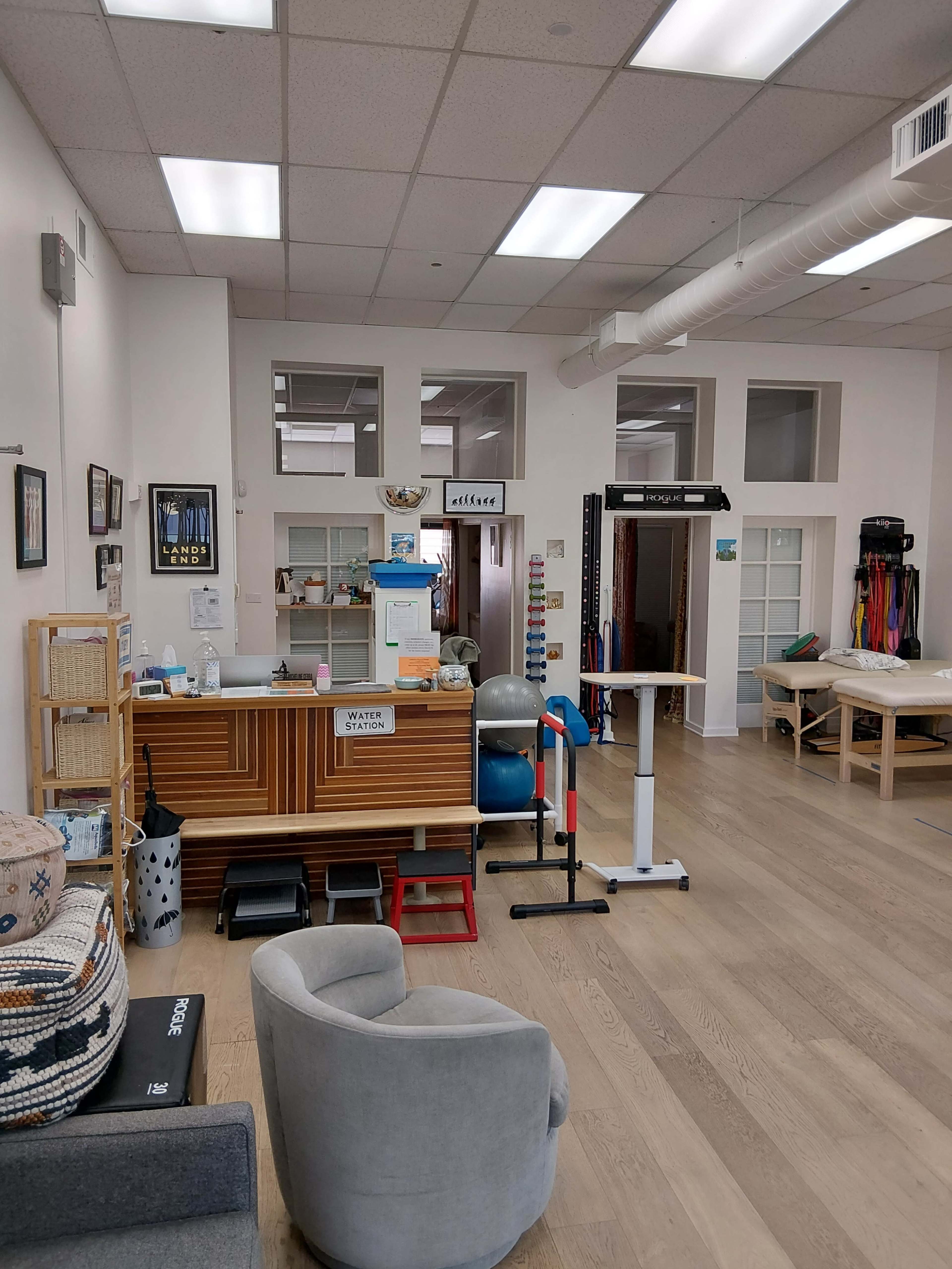 The image shows a bright, spacious therapy or rehabilitation room with various equipment and furniture arranged neatly, including a reception desk, exercise balls, and therapy tables.