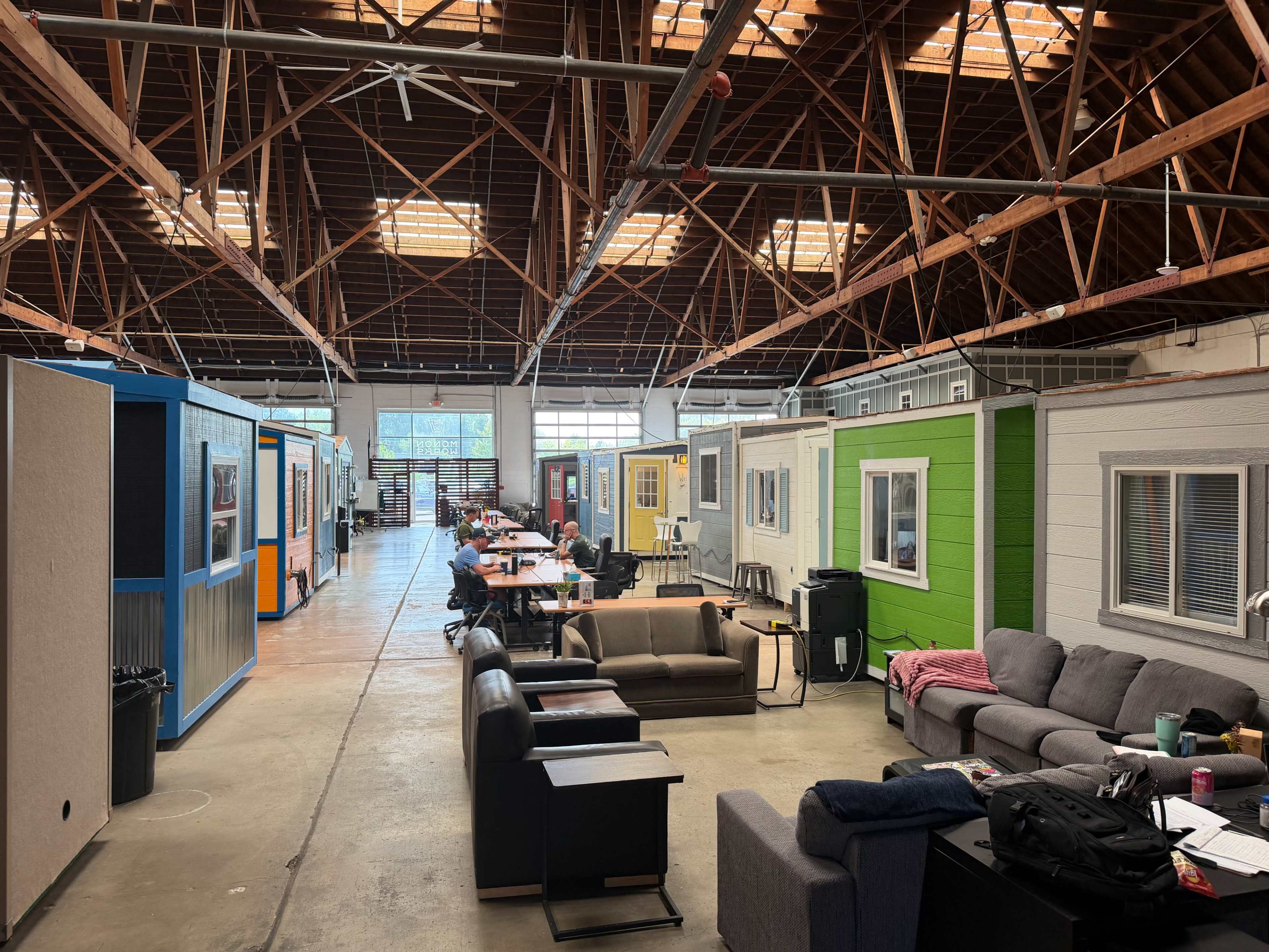 The image shows a large indoor space with several small cabins lined up along one side and tables for workstations in the center.