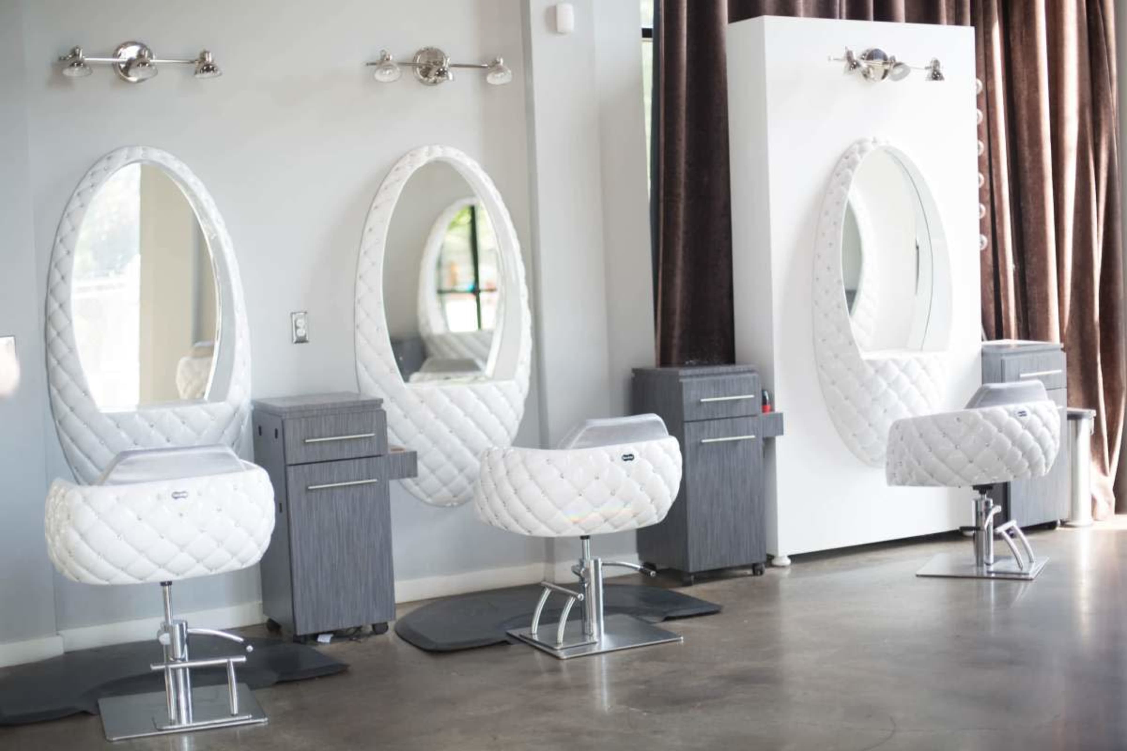 The image shows a modern hair salon with three oval mirrors and matching padded salon chairs, positioned against a neutral-colored wall and illuminated by overhead lights.