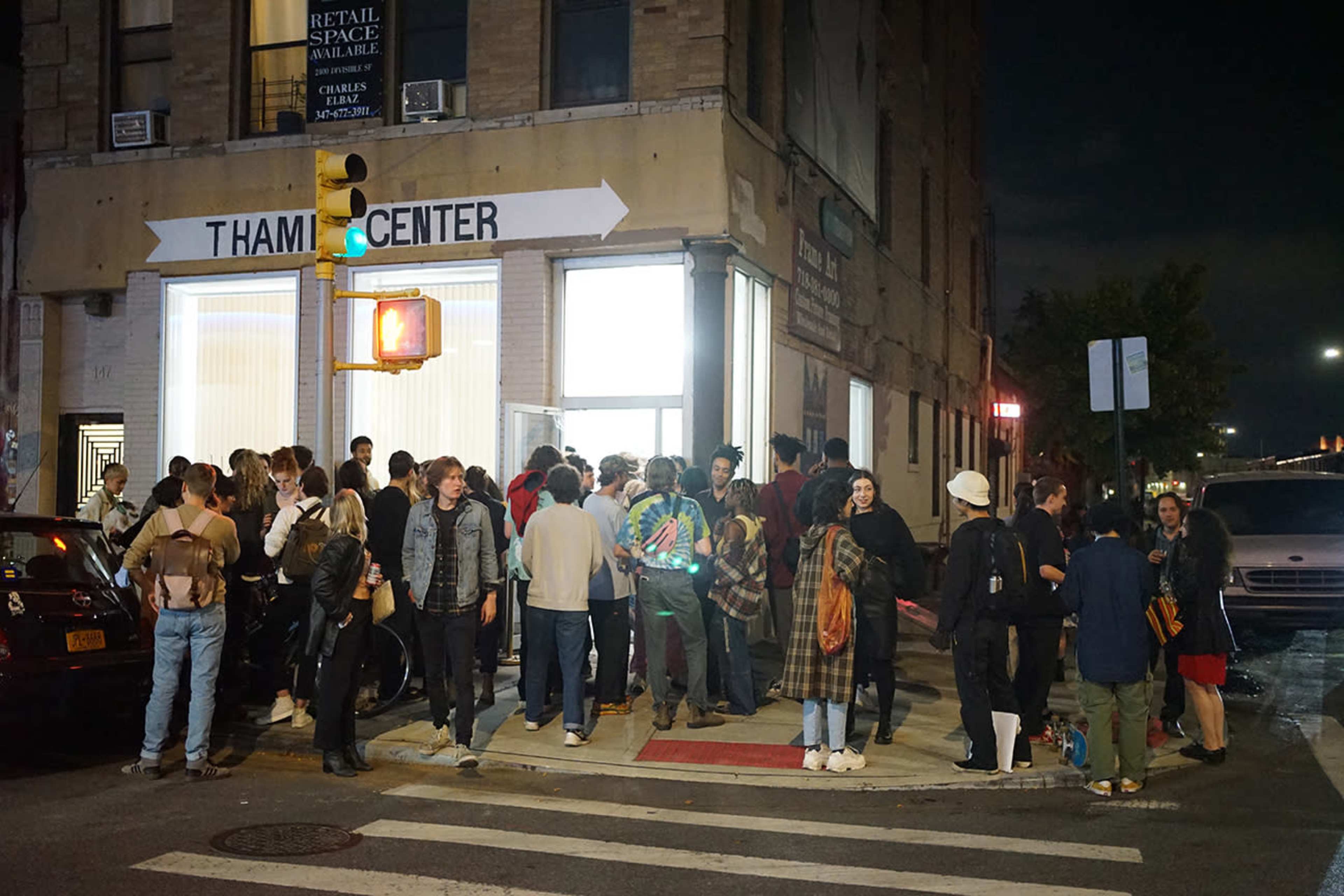 A large group of people gathers outside a building with a sign reading "THAM CENTER" on a busy urban corner at night.