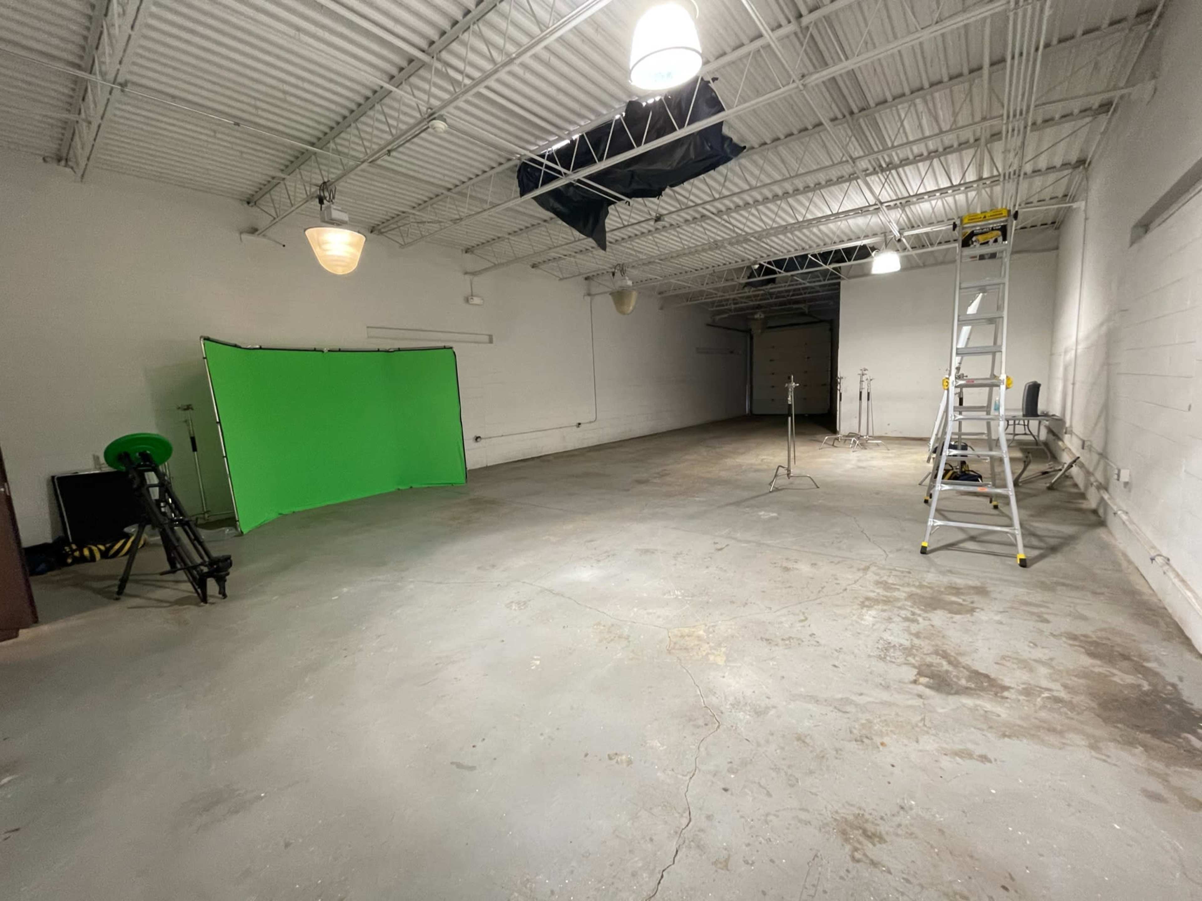 An empty warehouse space featuring a green screen setup, a ladder, and various lighting equipment.