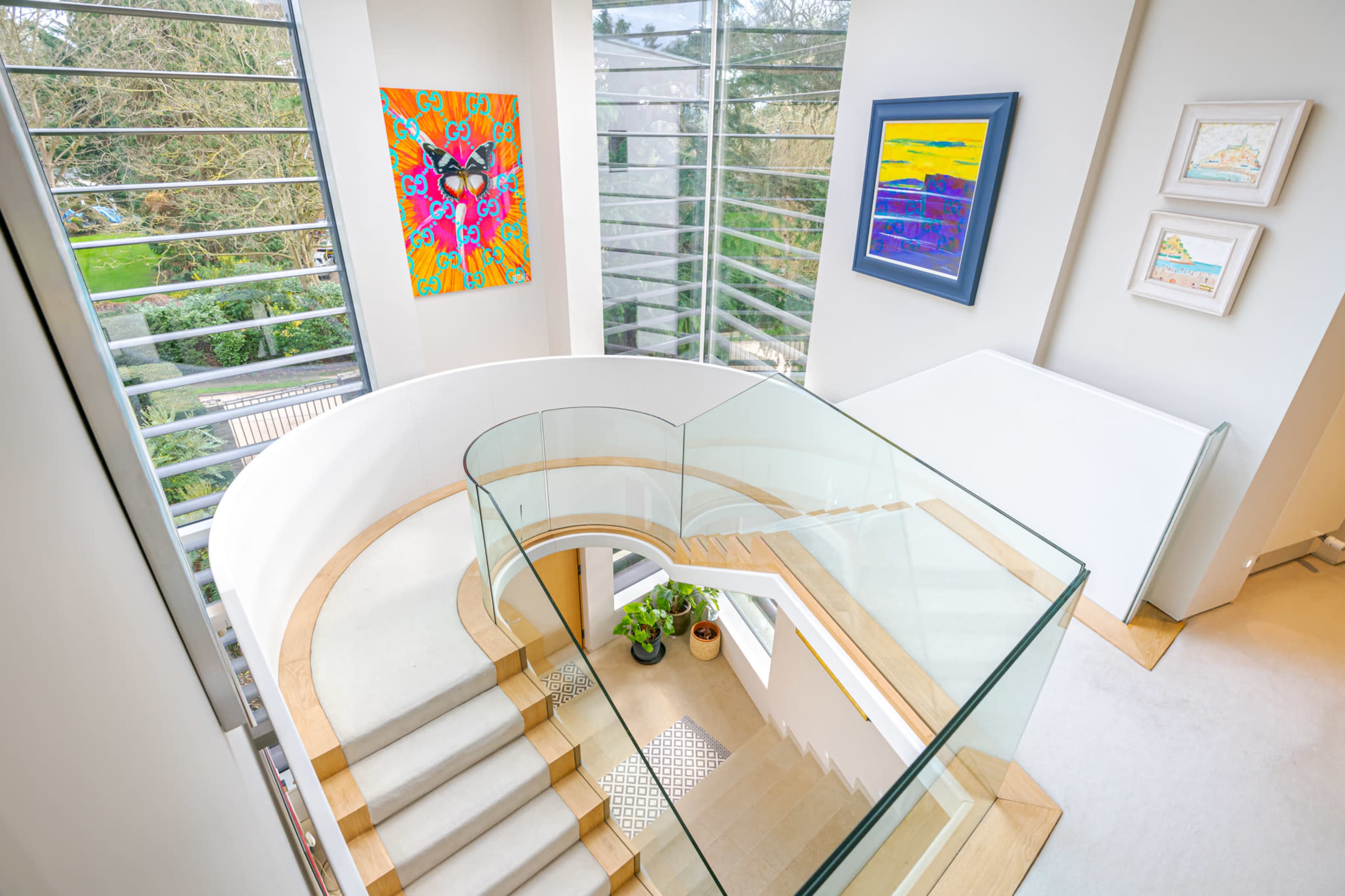 The image shows a modern, spiral staircase illuminated by natural light, with colorful artwork on the walls and greenery visible through large windows.