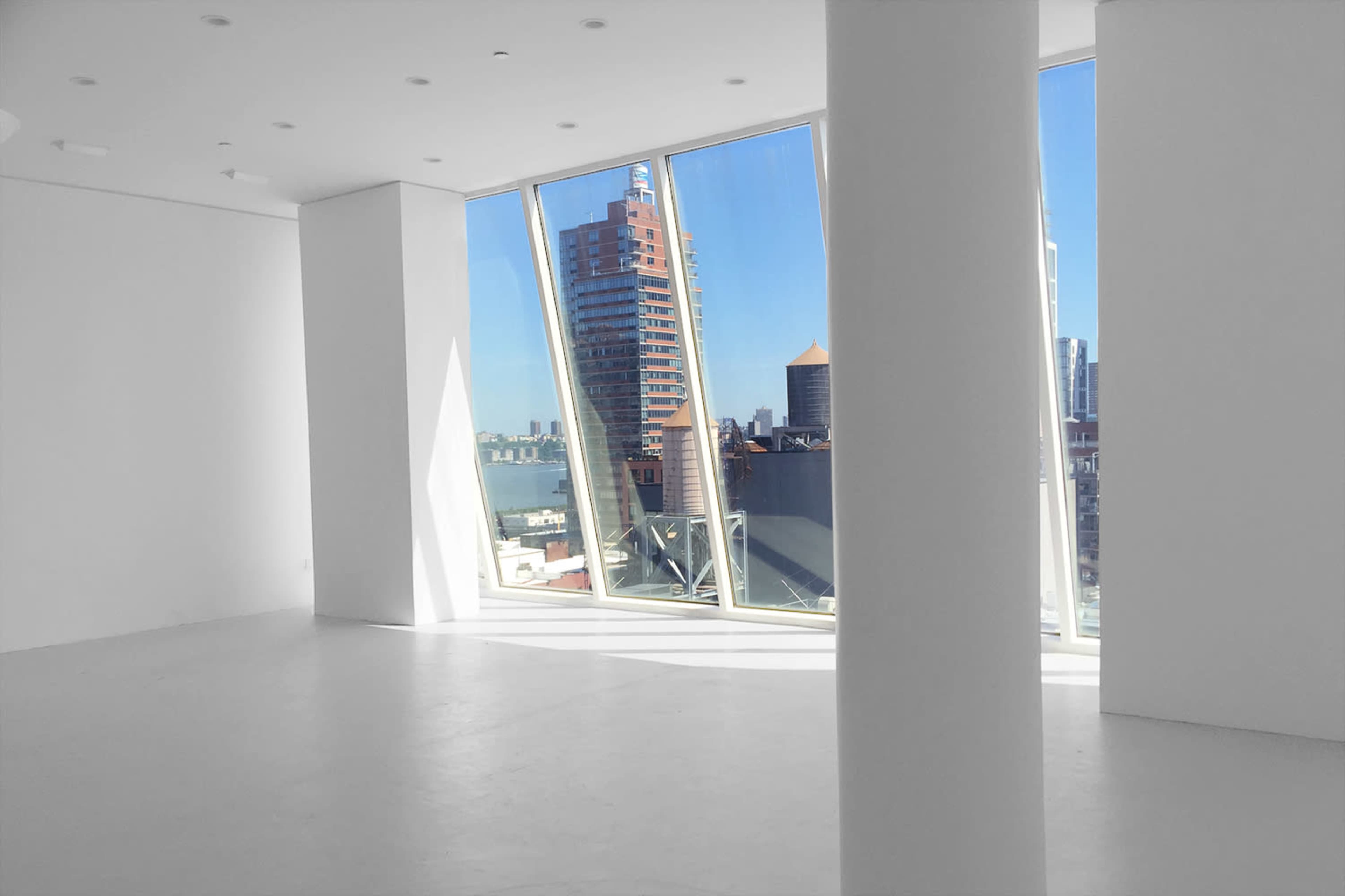 The image shows a spacious, modern room with large angled windows offering views of a city skyline and water.