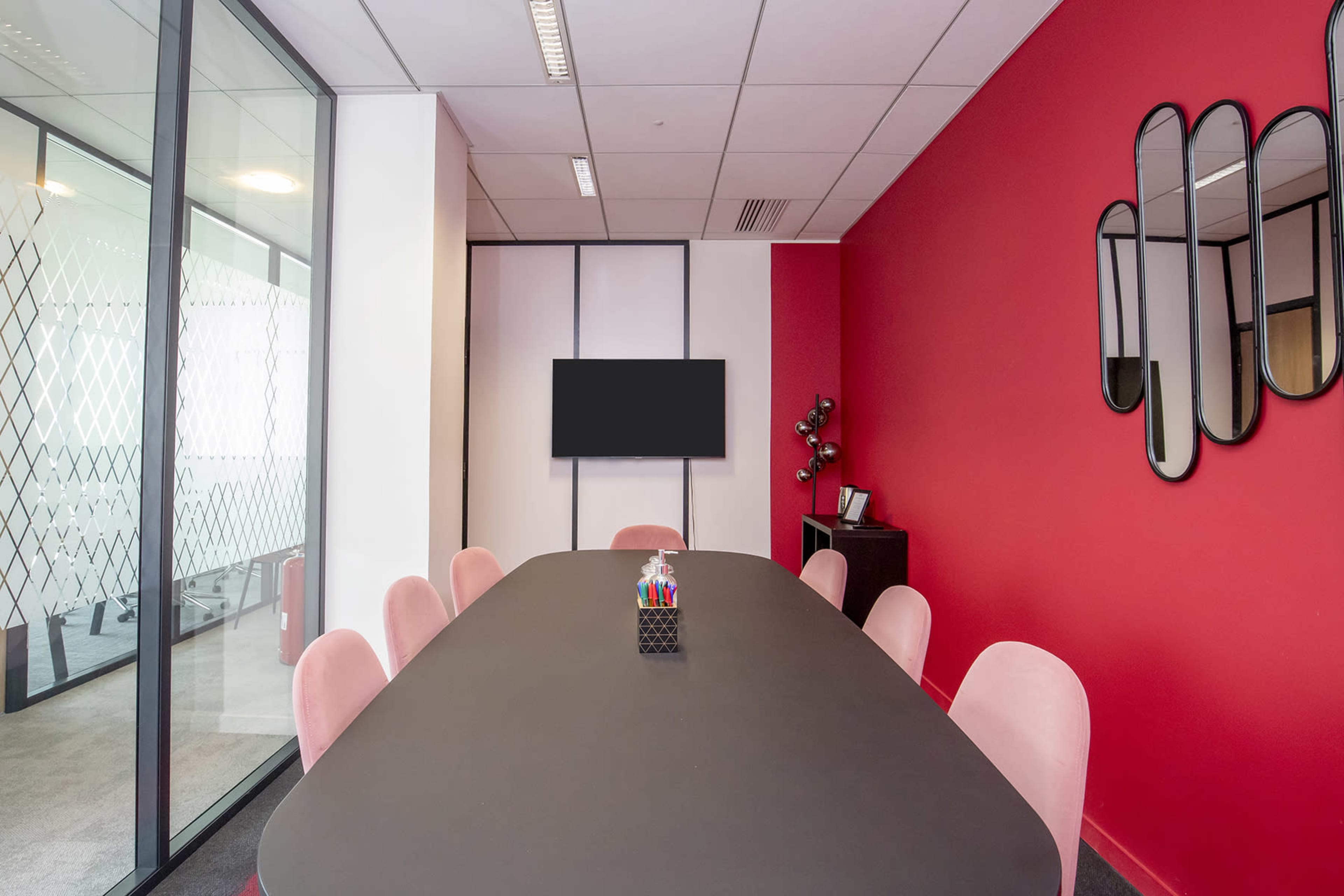 A modern conference room features a large black table surrounded by pink chairs, a wall-mounted screen, and decorative mirrors on the red wall.