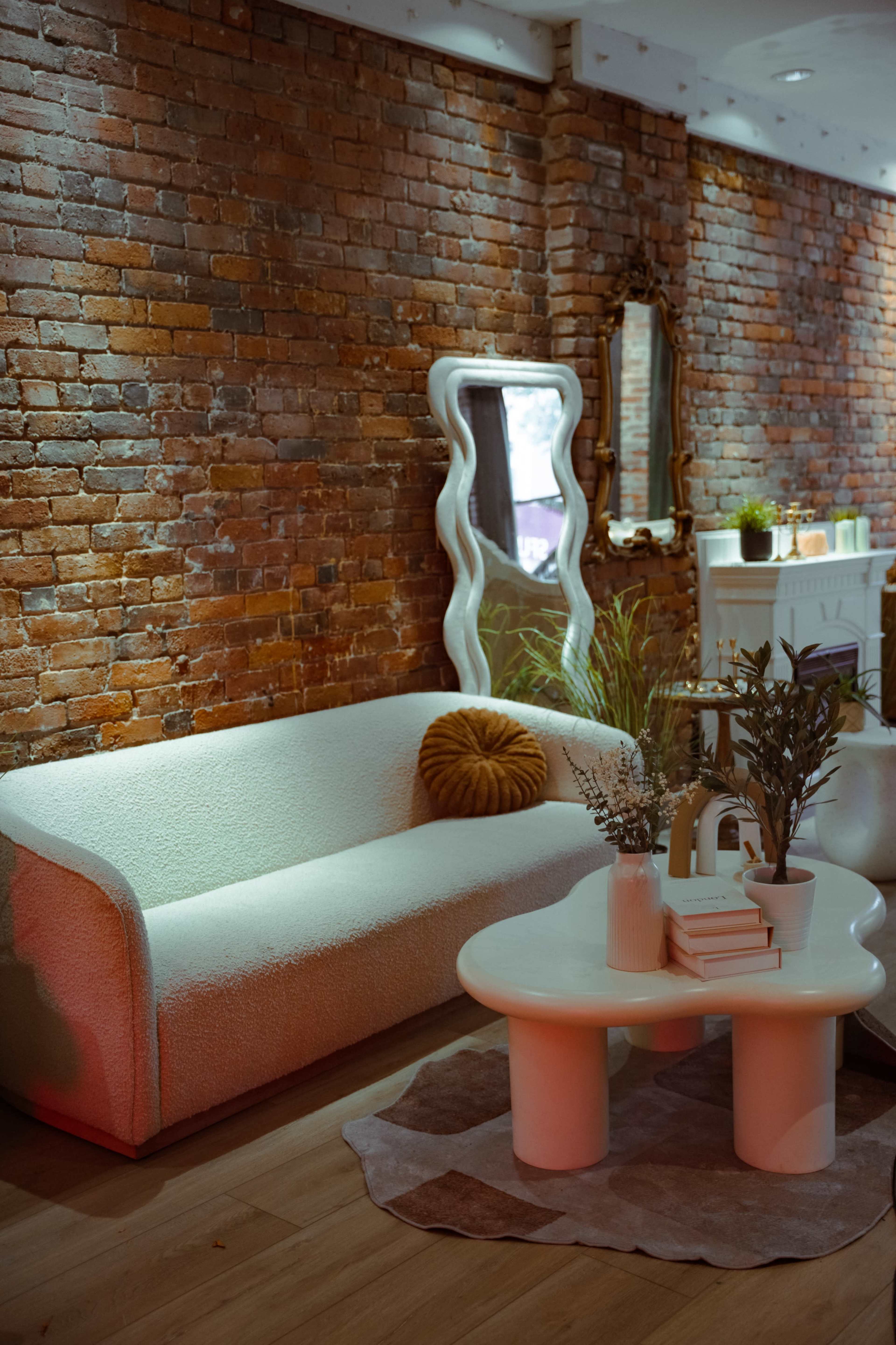 The image shows a cozy living room with a textured white sofa, a round white coffee table, and a mirror on a brick wall.