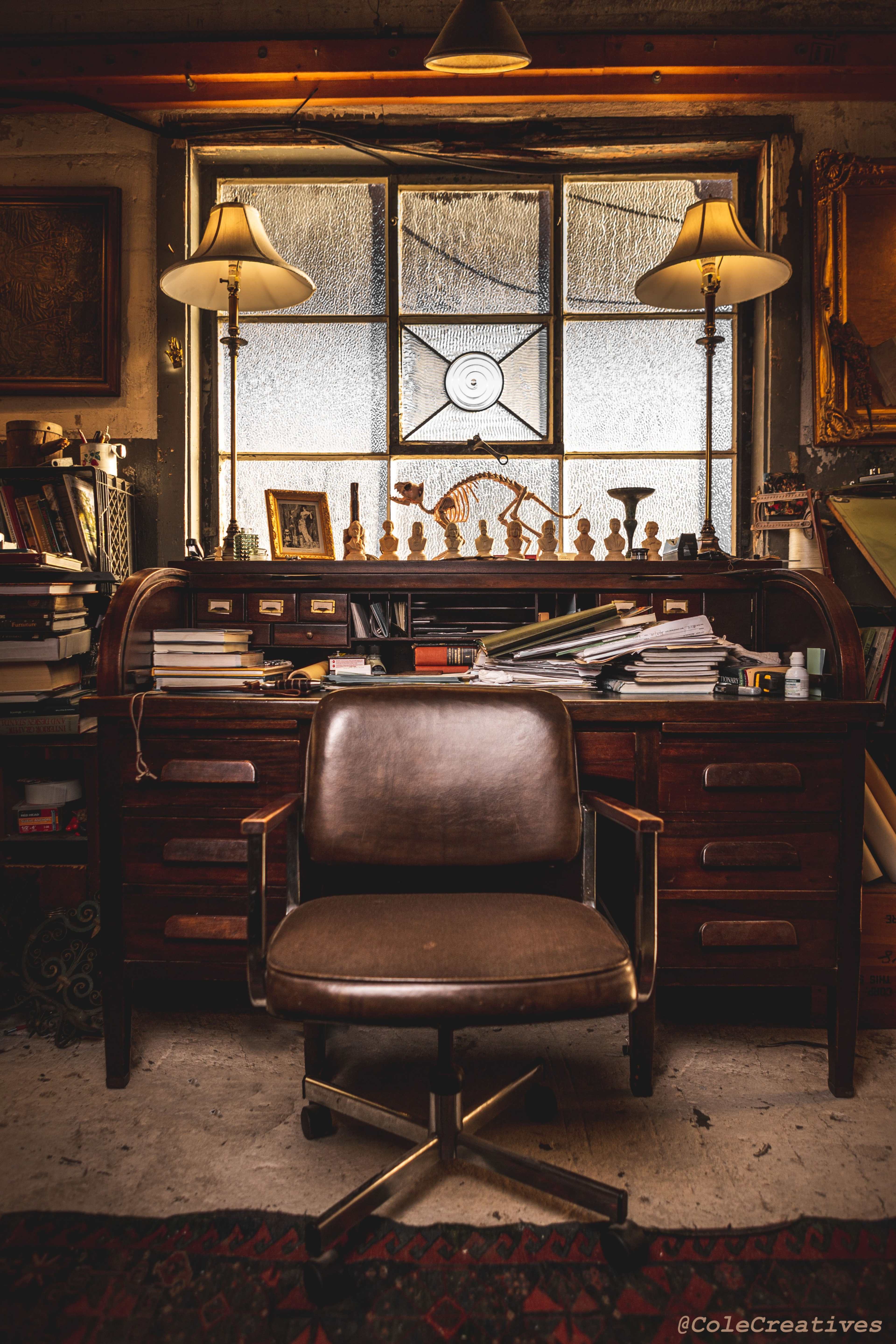 A wooden desk cluttered with books and papers stands in a dimly lit room, facing a brown rolling chair and a window adorned with decorative figurines.