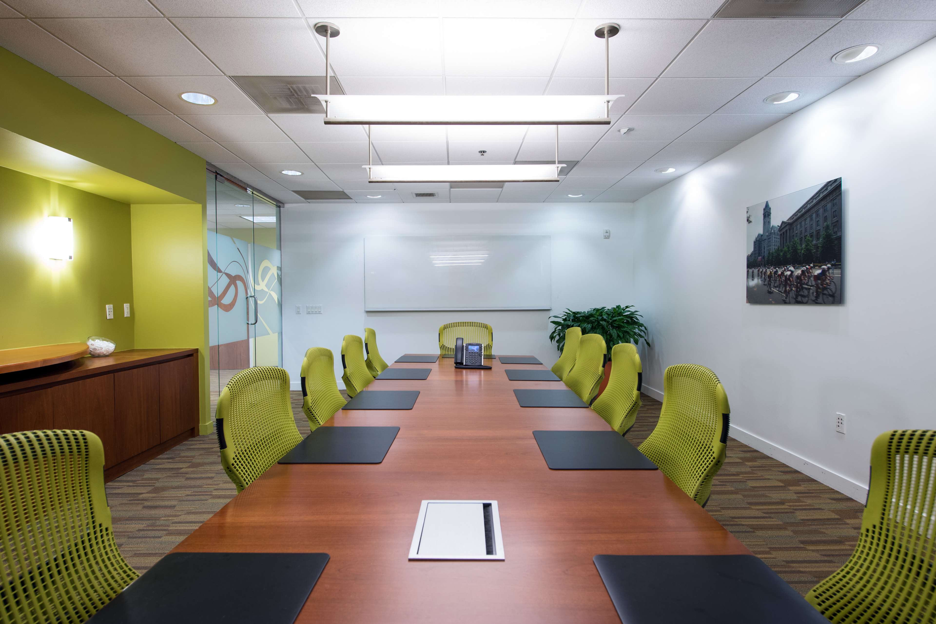 A modern conference room featuring a long wooden table with green ergonomic chairs, a whiteboard, and a large window with a view of a plant.
