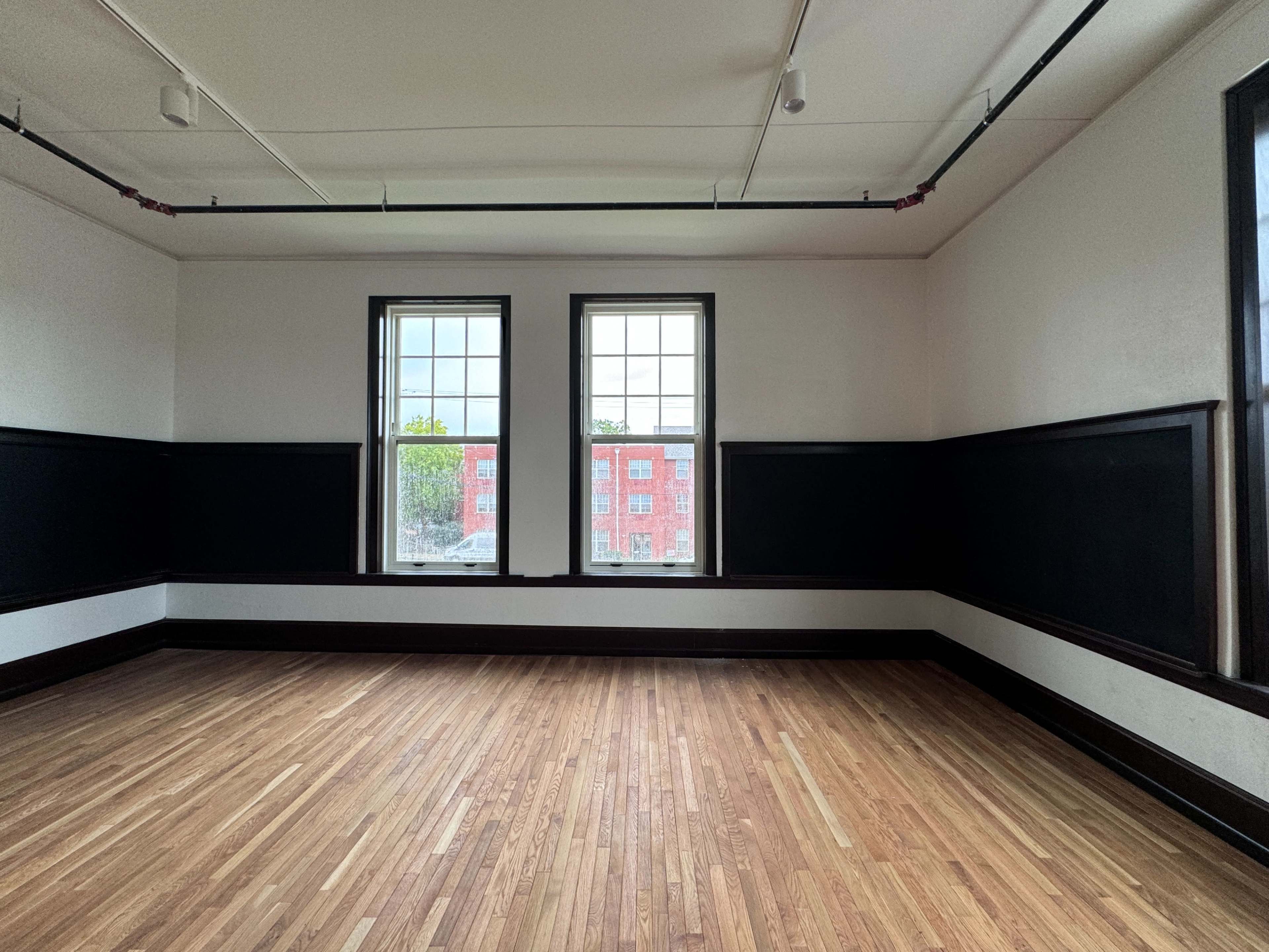 The image shows an empty room with hardwood floors, two windows, and dark-painted walls that form a wainscoting design.