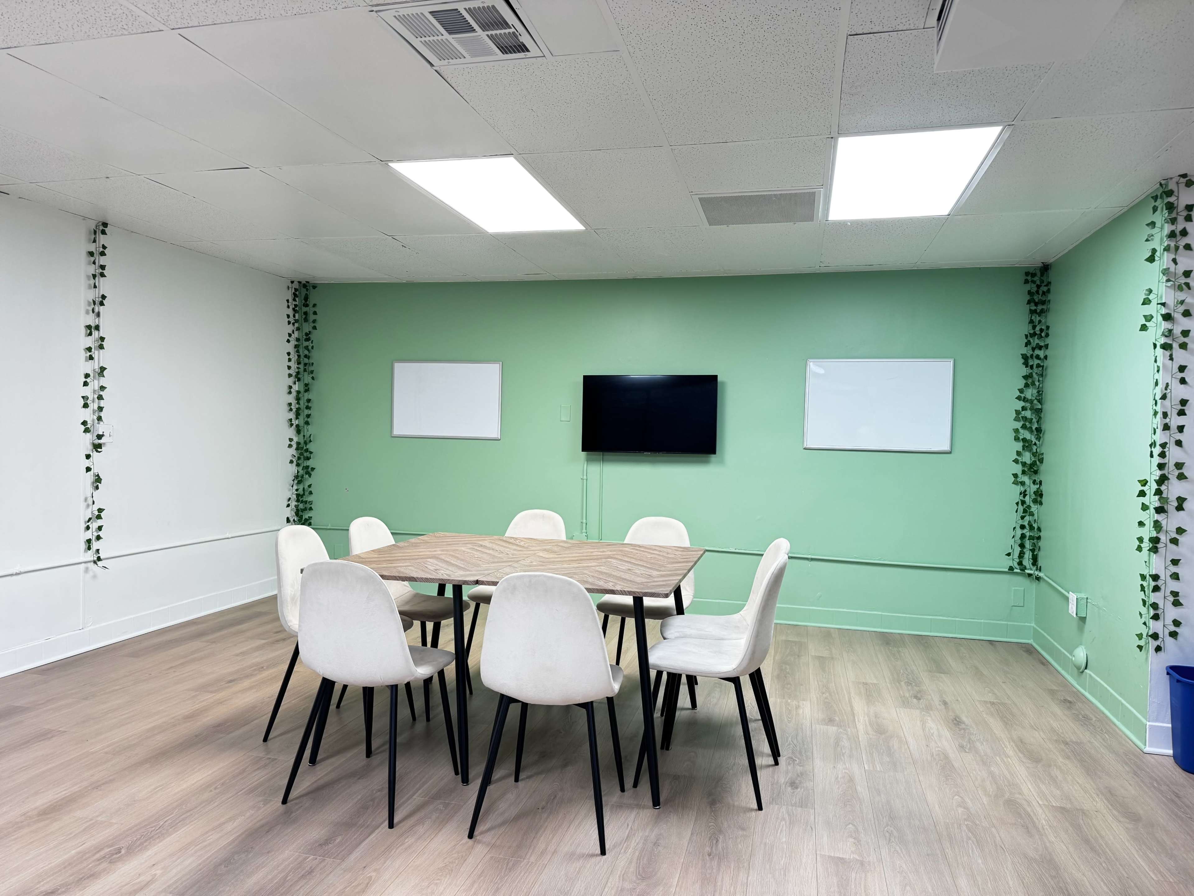 The image shows a meeting room with a wooden table surrounded by six white chairs, green walls, and a television mounted on the wall, along with two whiteboards.