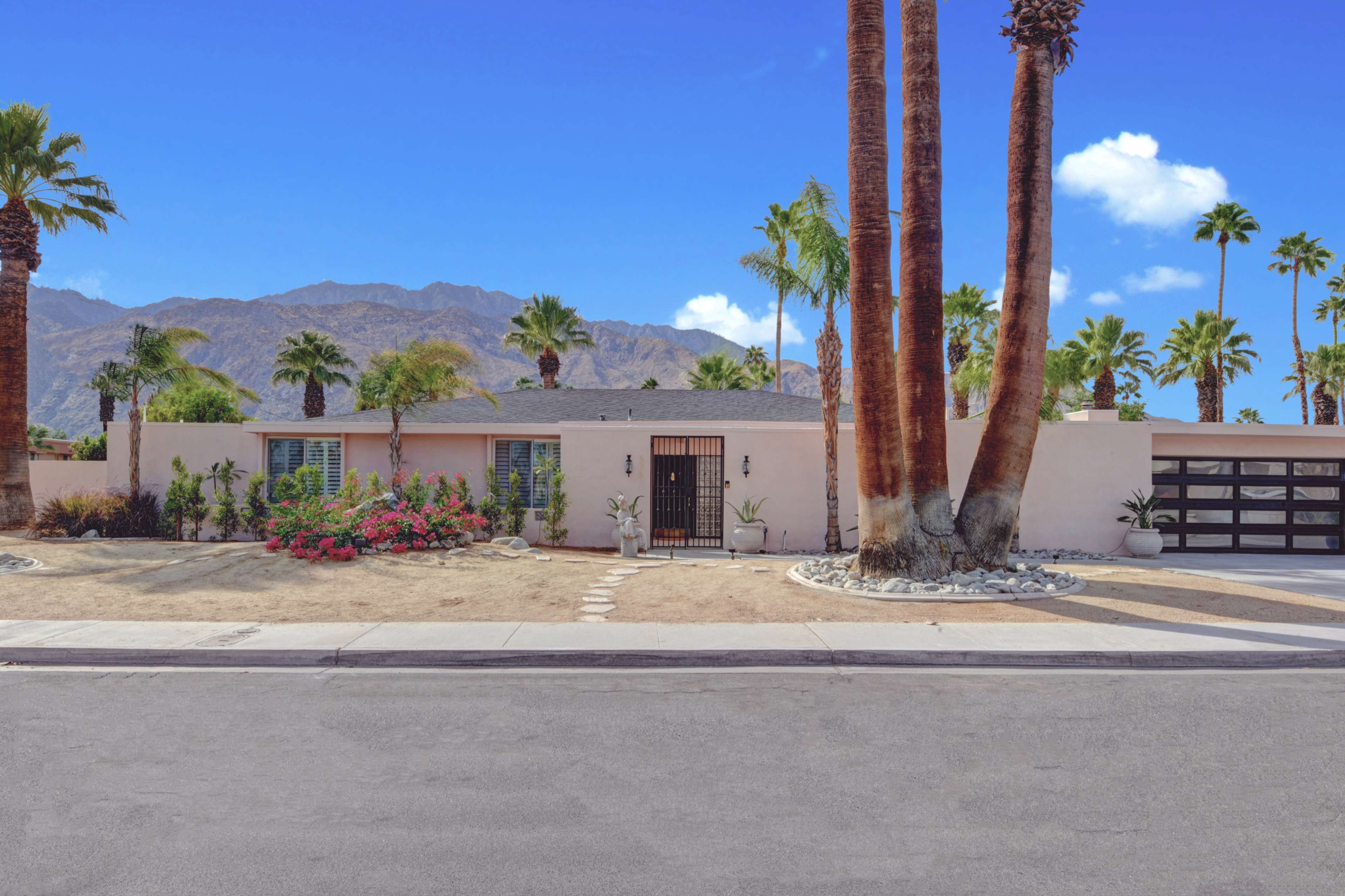 A single-story house with a decorative front gate, surrounded by palm trees and low landscaping, sits against a backdrop of mountains.