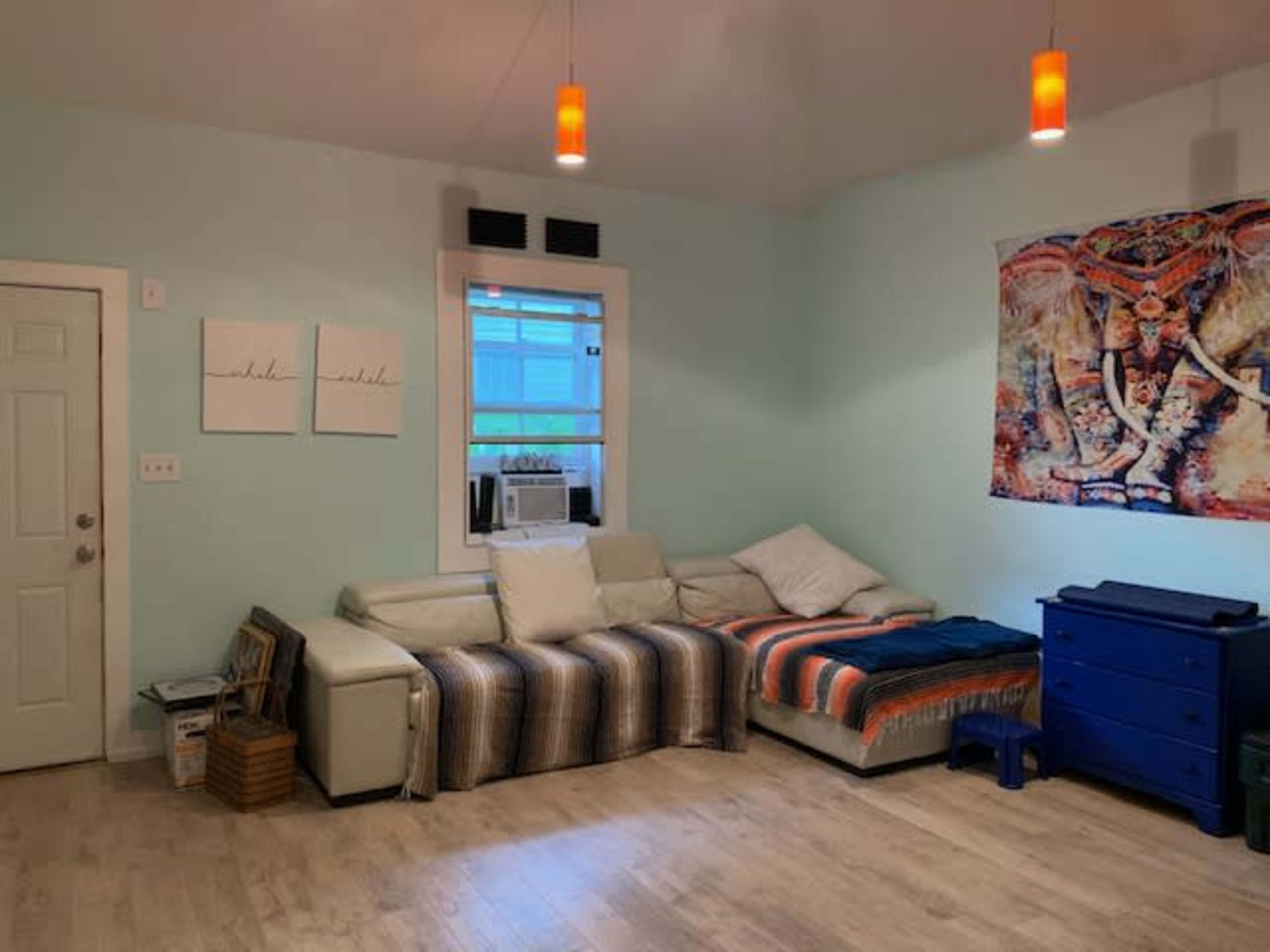 A light blue room with a beige sectional sofa, a bed with colorful bedding, a blue dresser, and decorative artwork on the wall.