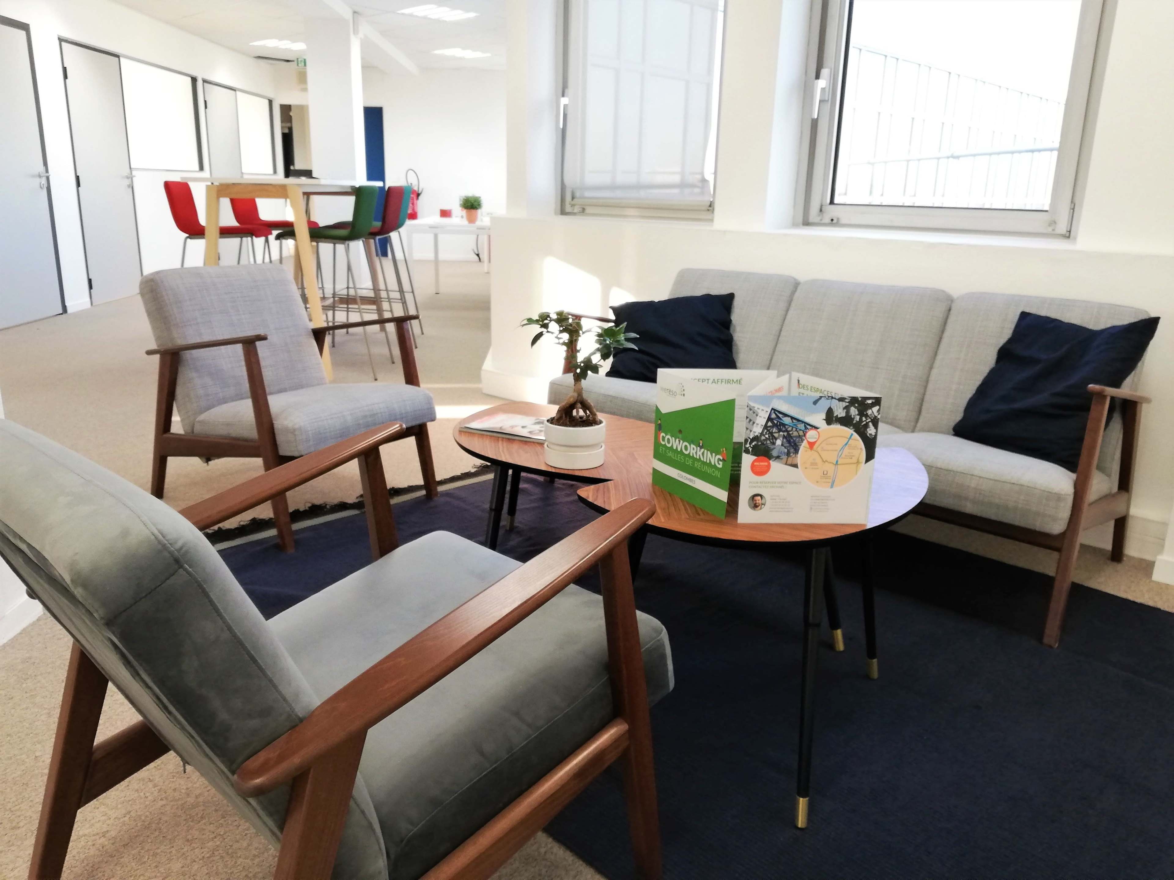 A modern office lounge area with two armchairs, a small round table displaying a brochure, and a sofa, surrounded by bright windows and light-colored walls.
