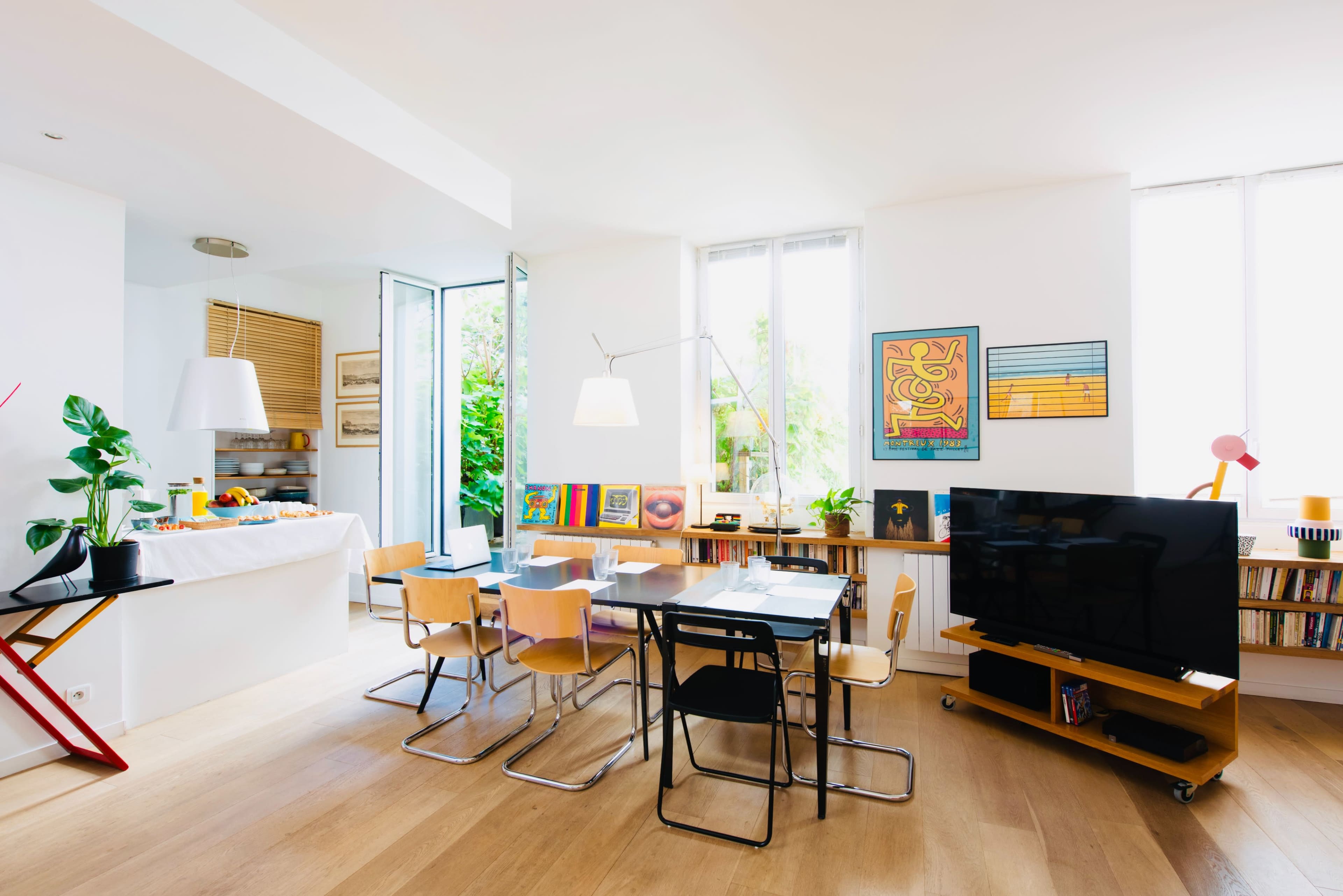 A bright, open living space features a dining area with a table and chairs, a modern TV stand, and large windows leading to a garden.
