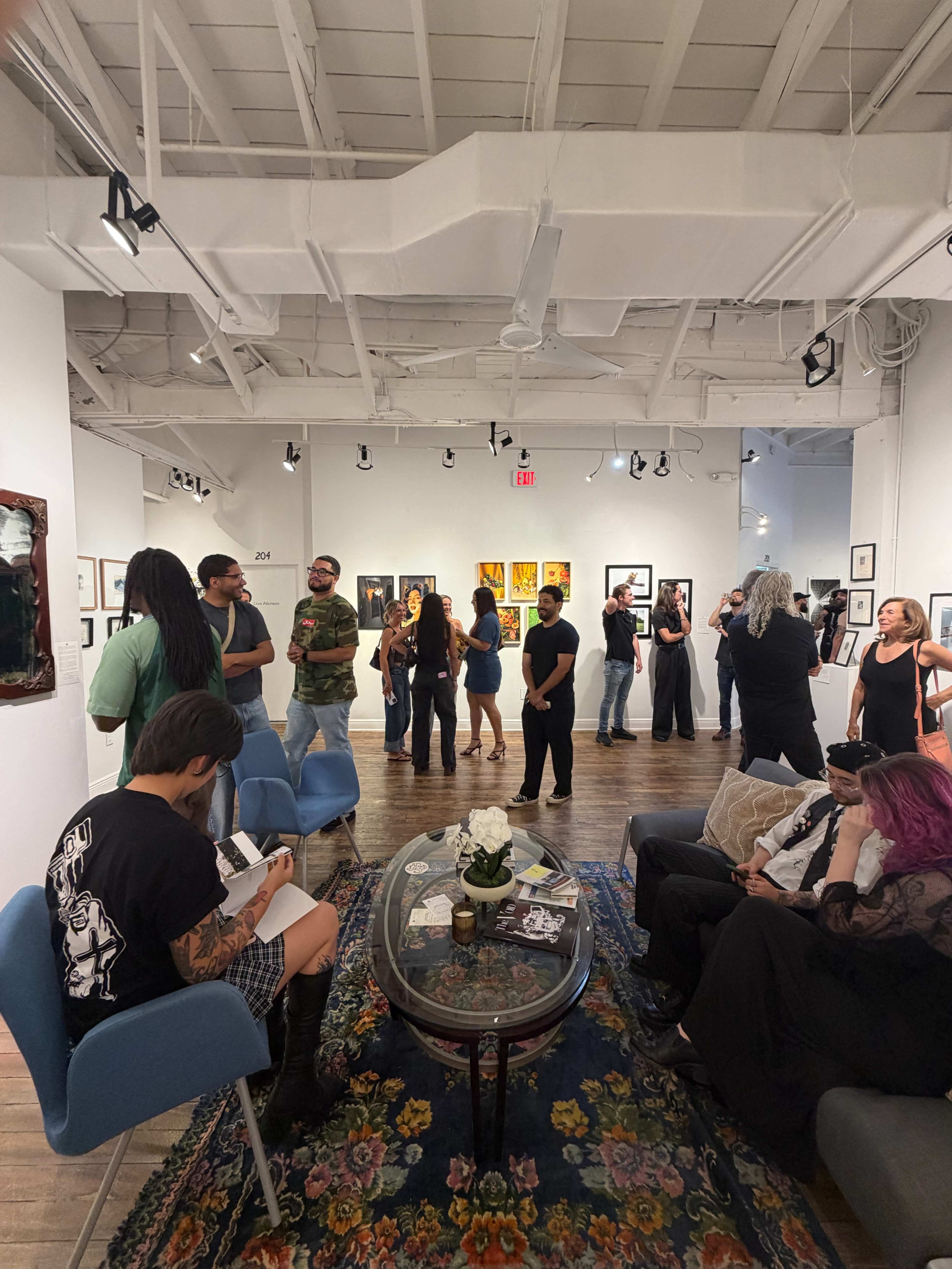 The image shows people mingling in an art gallery featuring various artworks on the walls, with some attendees seated on blue chairs and others standing in groups.