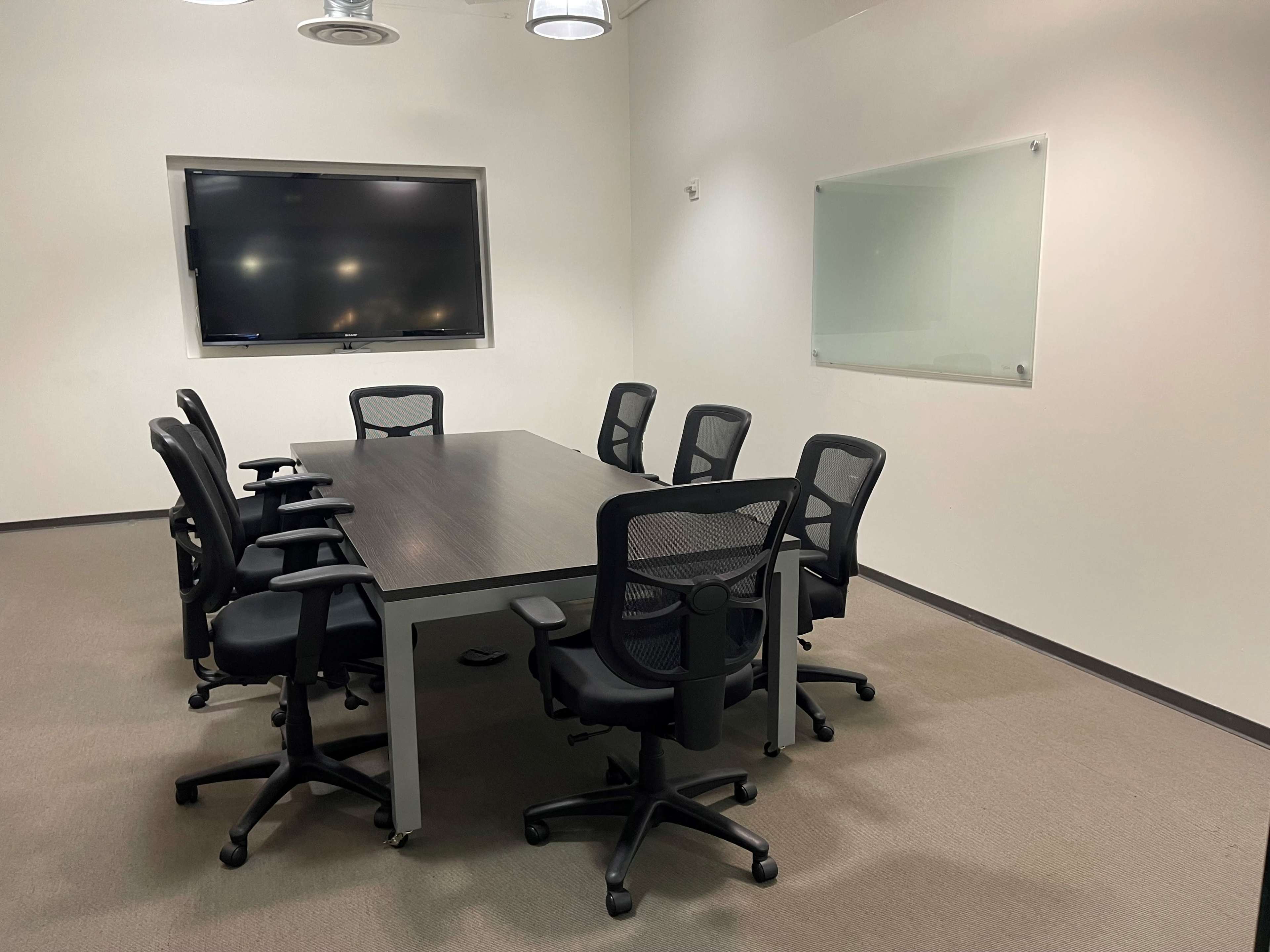 A conference room with a large table surrounded by eight black office chairs and a wall-mounted screen.
