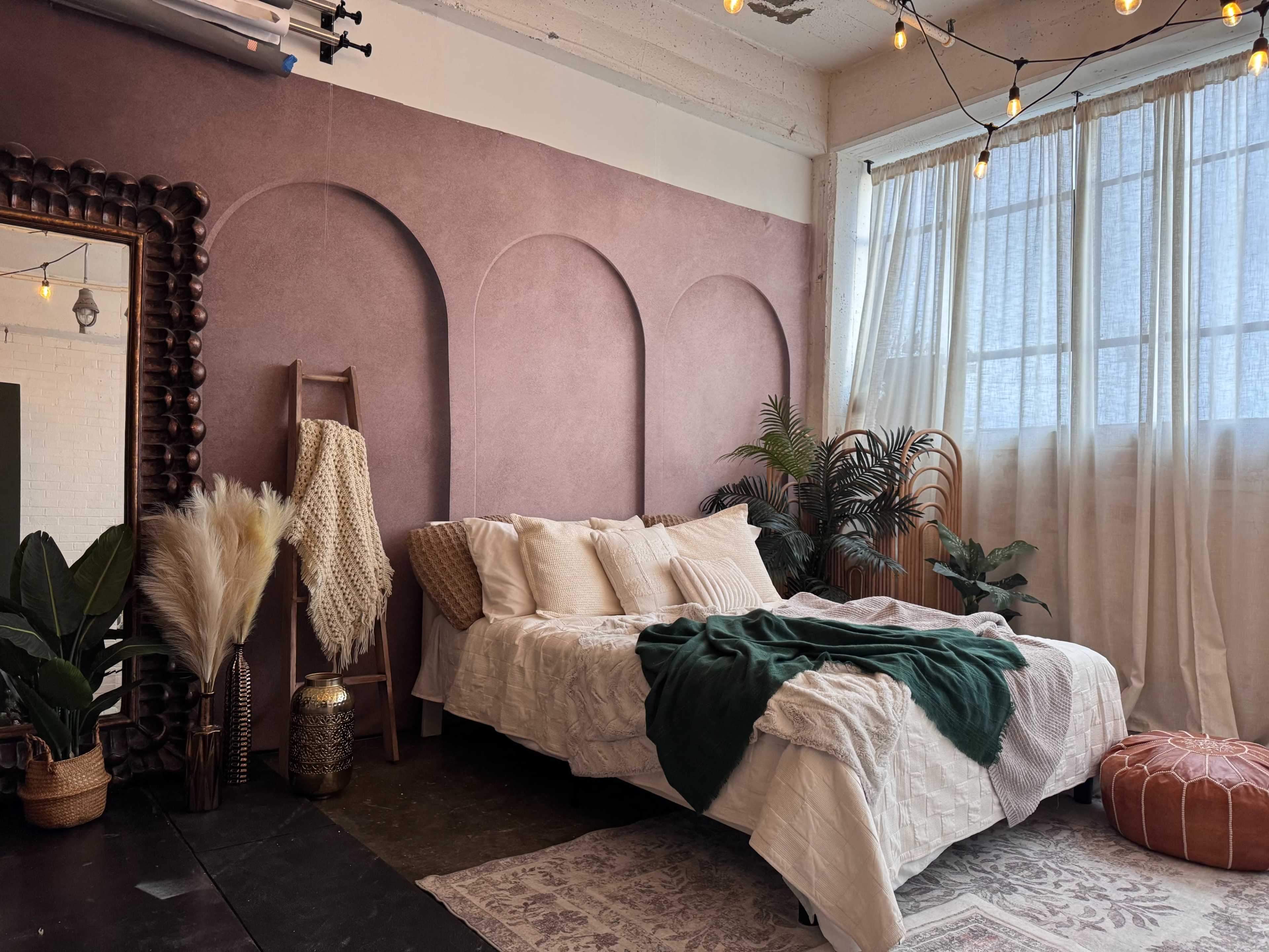 The image shows a decorated bedroom featuring a bed with cream-colored bedding, a large mirror, plants, and string lights hanging above.