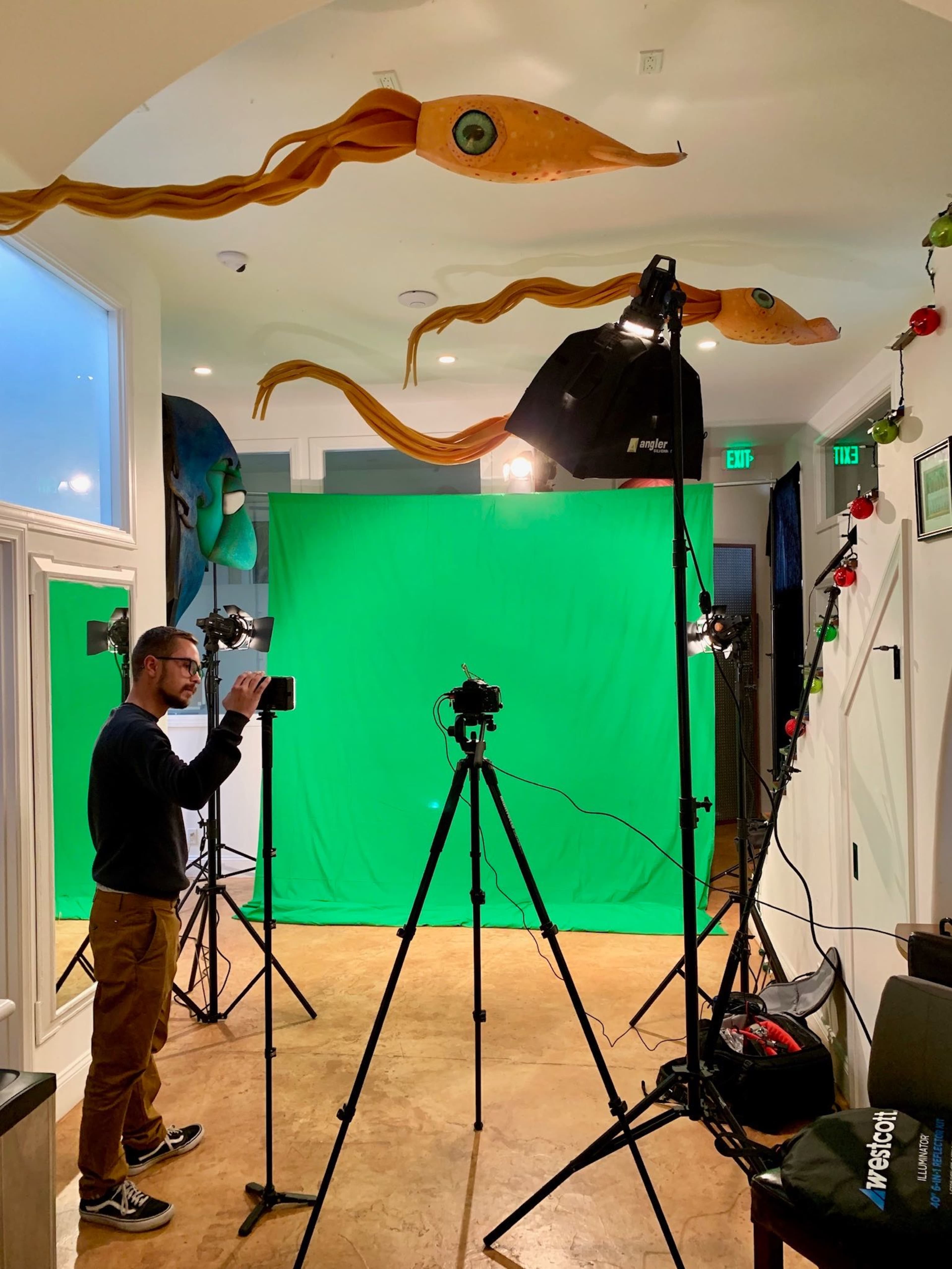 A man sets up a camera in front of a green screen, surrounded by studio lighting and equipment in a recording space.