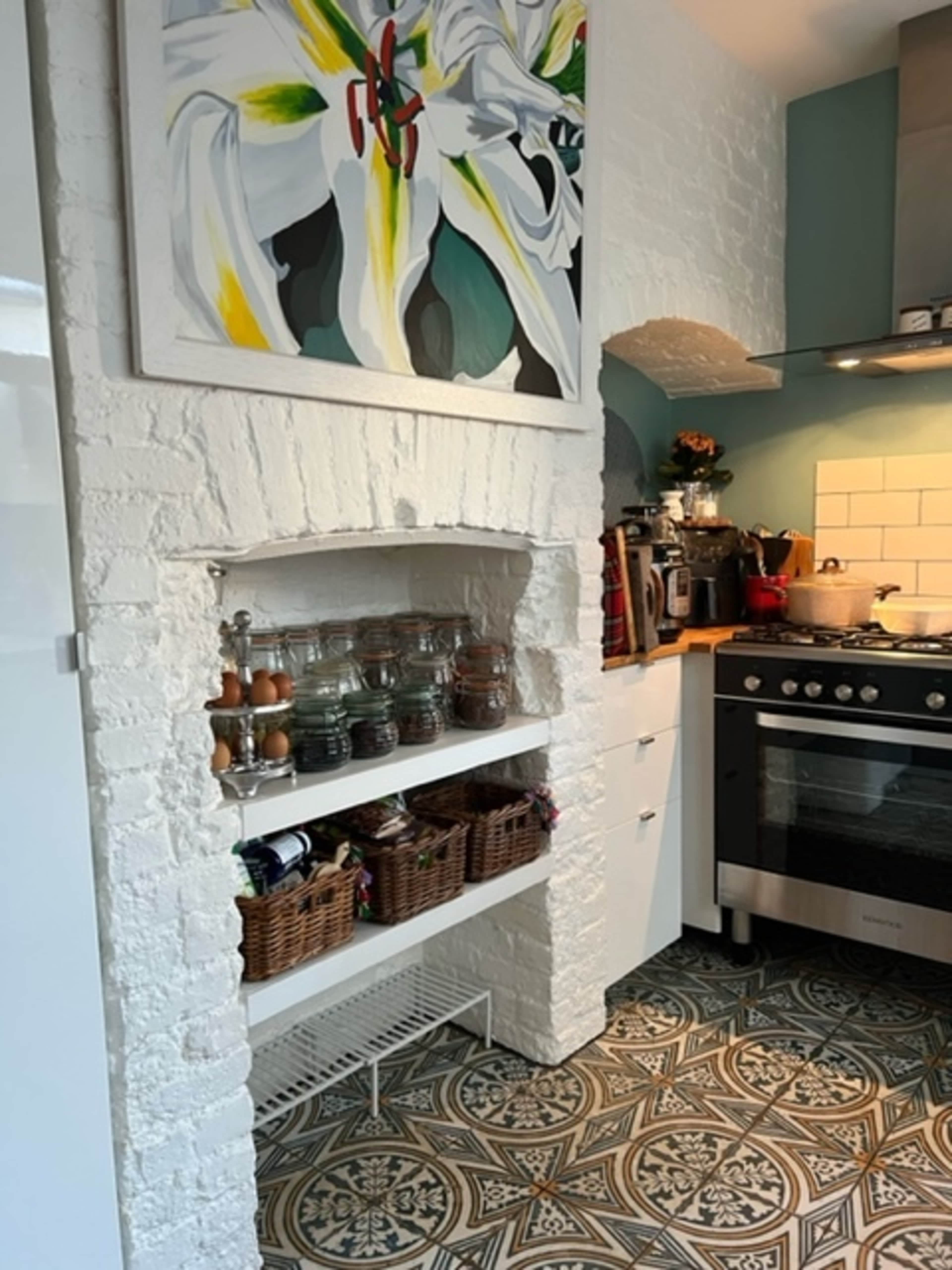 A kitchen with a white brick archway housing glass jars and baskets, a gas stove, and a colorful painting on the wall.
