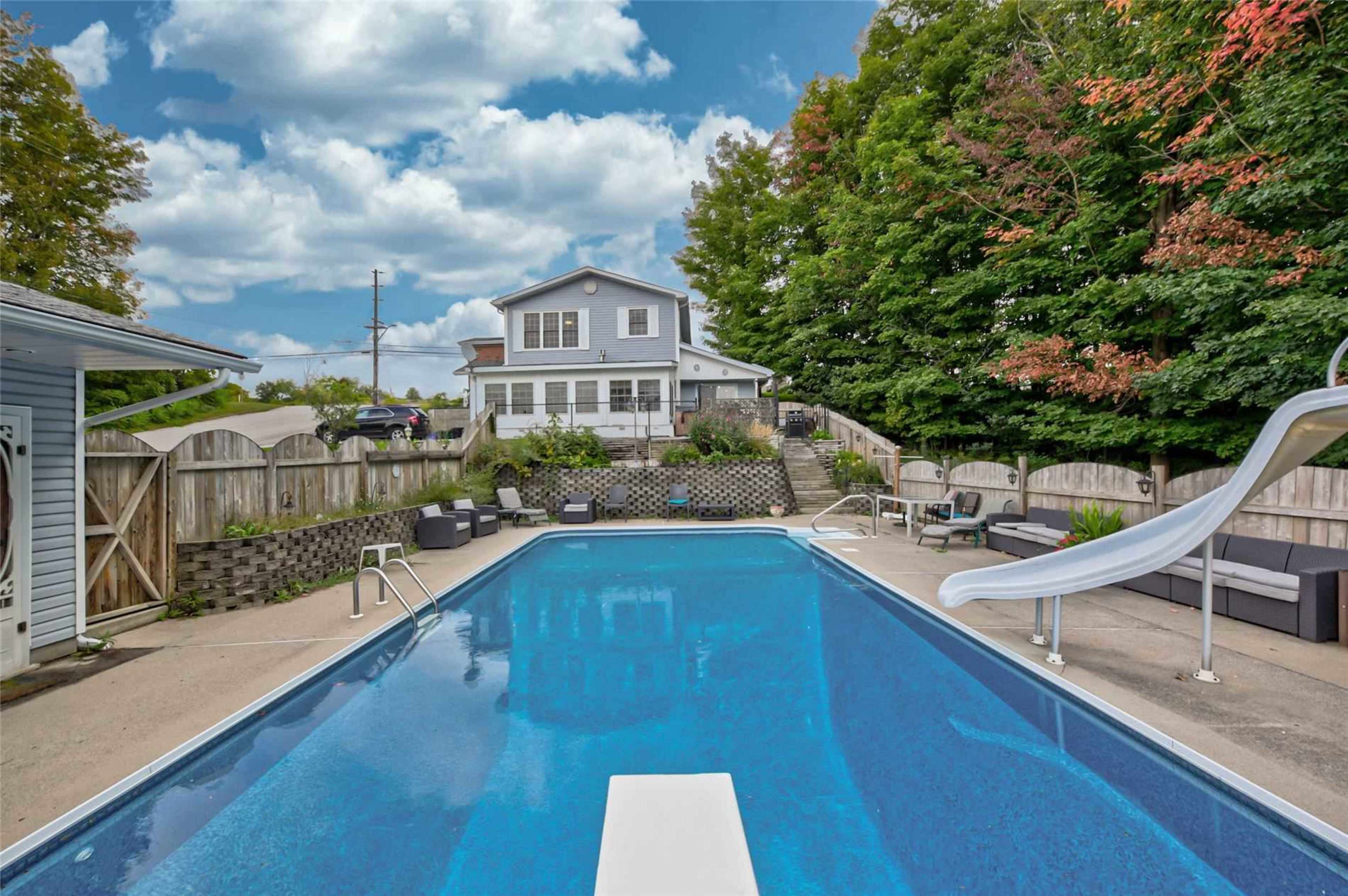A swimming pool with a diving board and a waterslide, surrounded by a patio area and landscaped greenery adjacent to a two-story house.