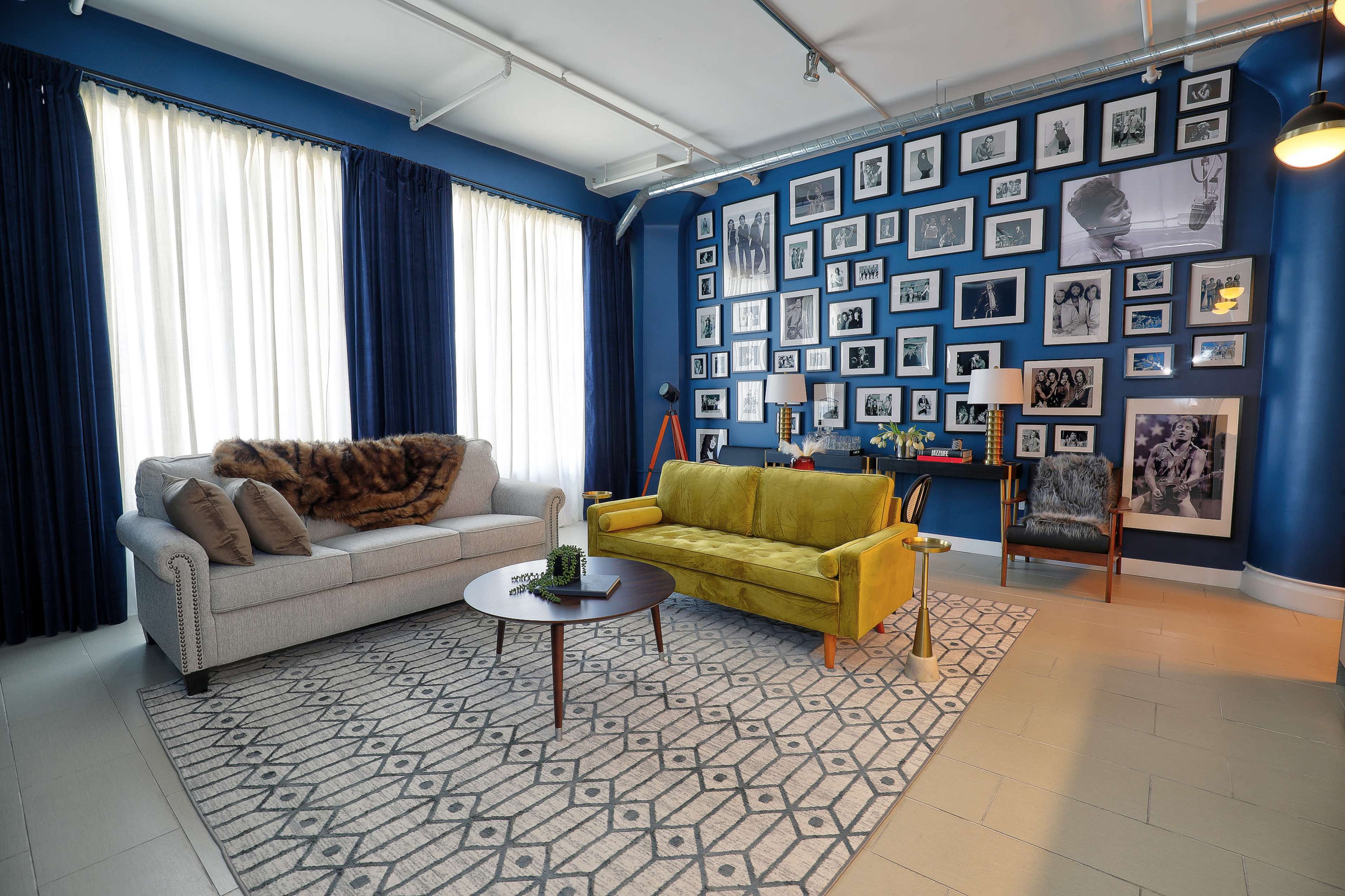 The living room features a pair of sofas, one beige with pillows and a fur throw, and one yellow, set against a blue wall adorned with a gallery of framed black-and-white photos.