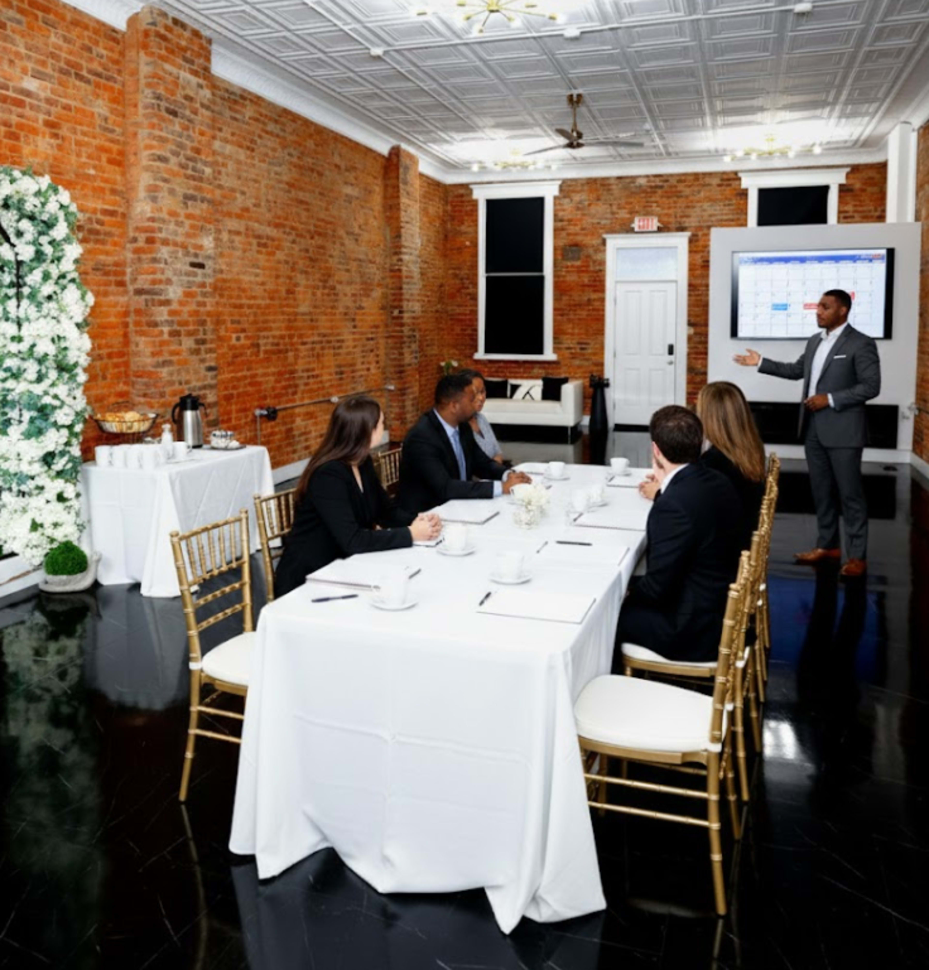 A speaker presents to a group seated at a conference table in a modern meeting room with exposed brick walls and a large screen displaying a presentation.