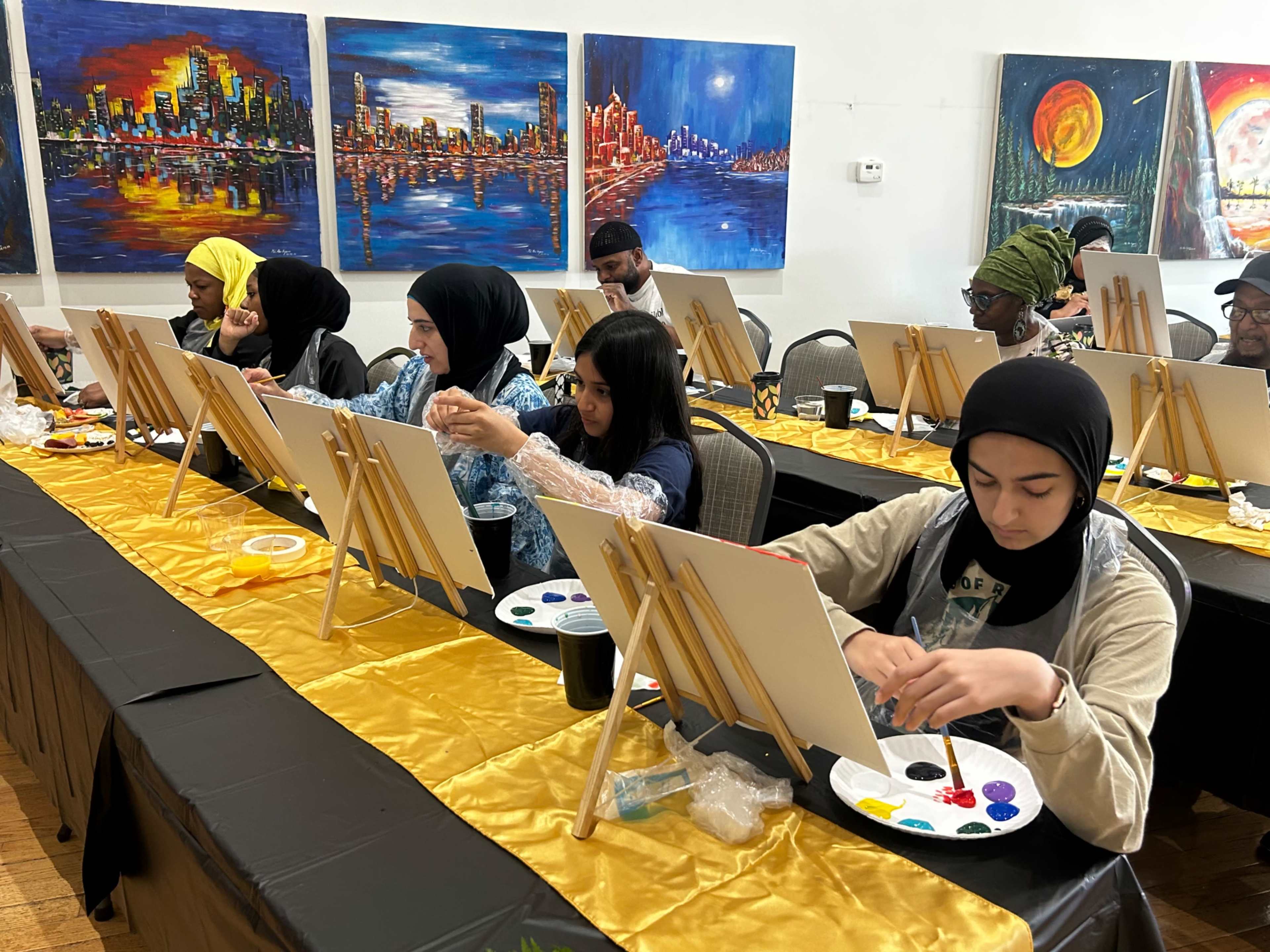 A group of individuals, dressed in various styles, is seated at a long table with blank canvases and paint palettes, engaged in a painting activity.