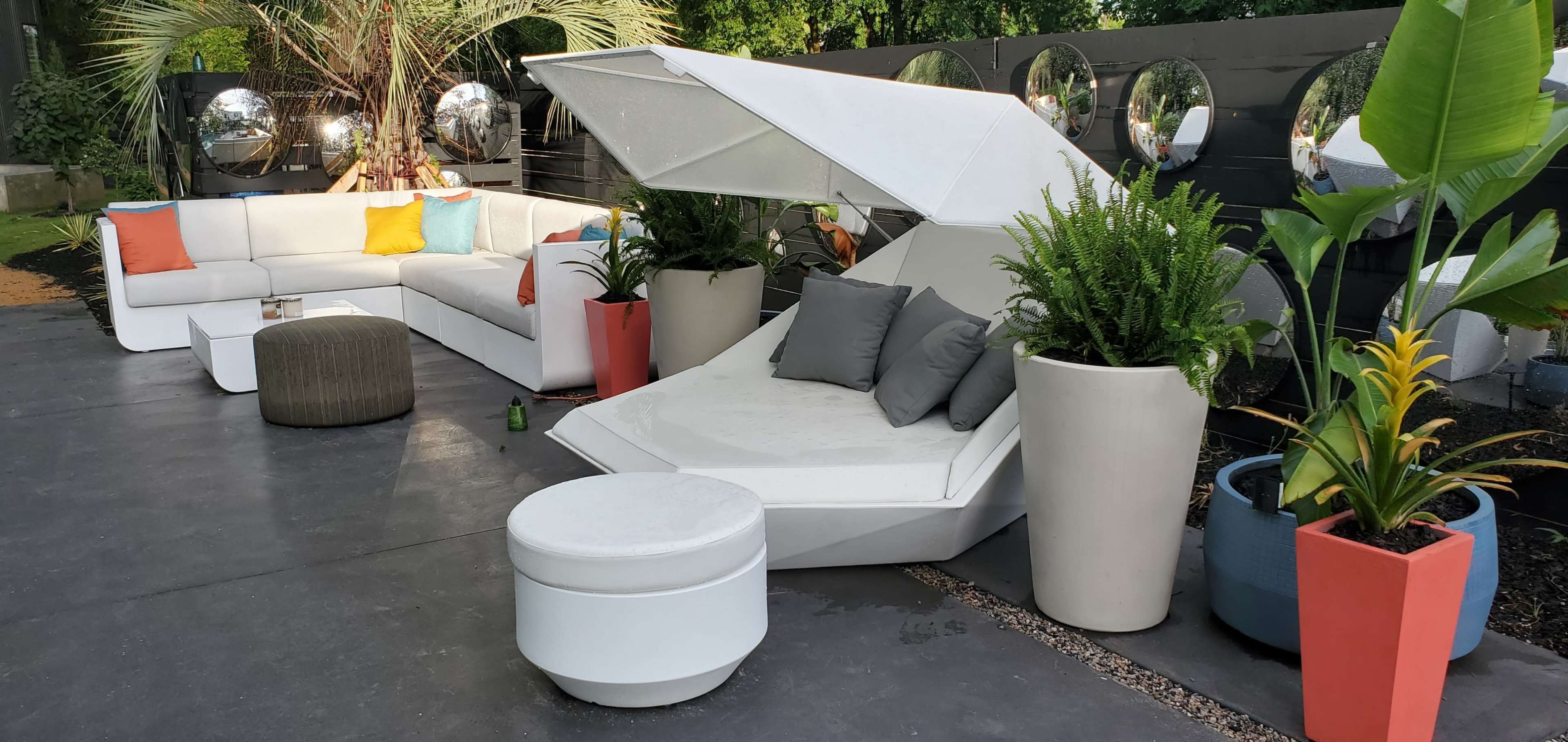 The image shows a modern outdoor seating area featuring a large white couch, a sleek lounging chair with an umbrella, and various potted plants arranged on a stone patio.