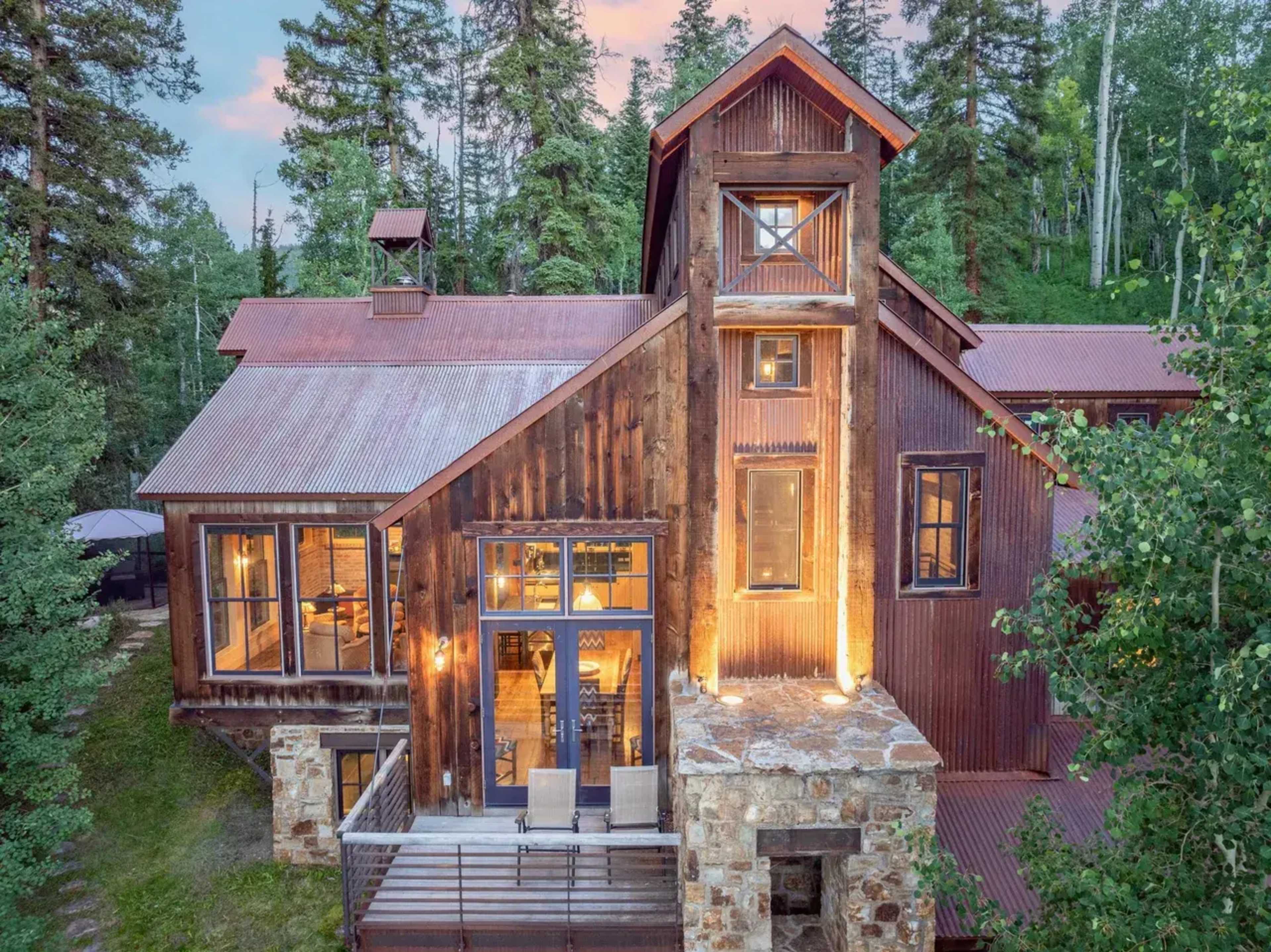 The image shows a rustic wooden house with a stone base, set among tall trees and illuminated by exterior lights during twilight.
