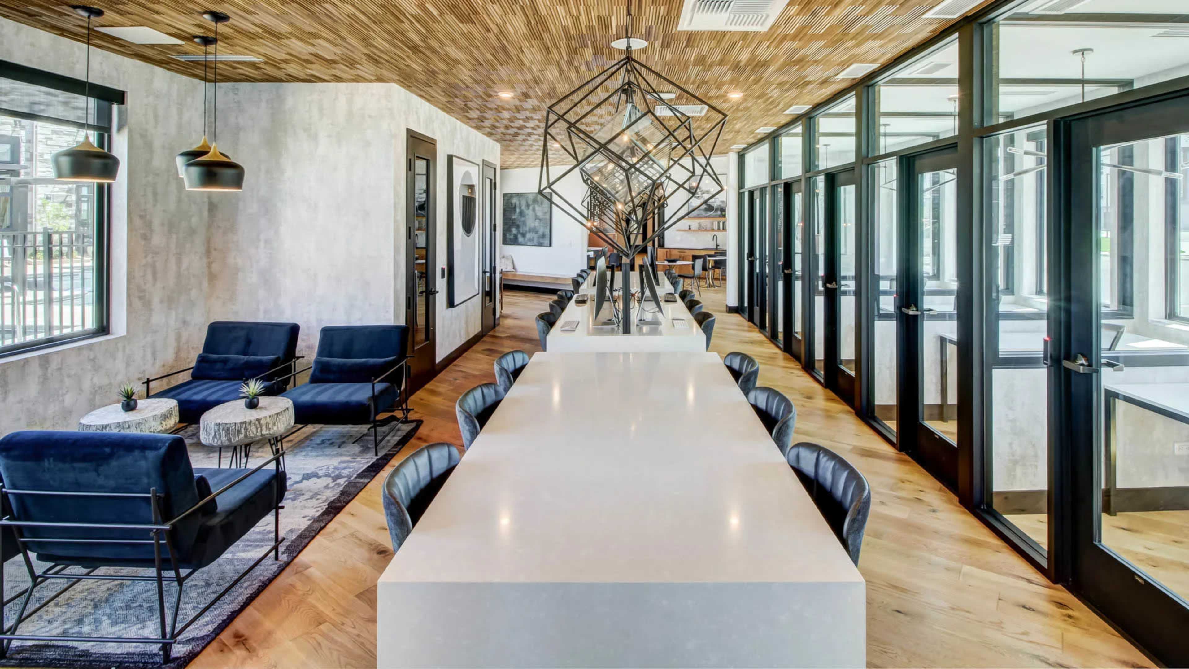 A modern office space featuring a long white table with black chairs, surrounded by seating areas with blue upholstered chairs and decorative tables, under a wooden ceiling and large windows.