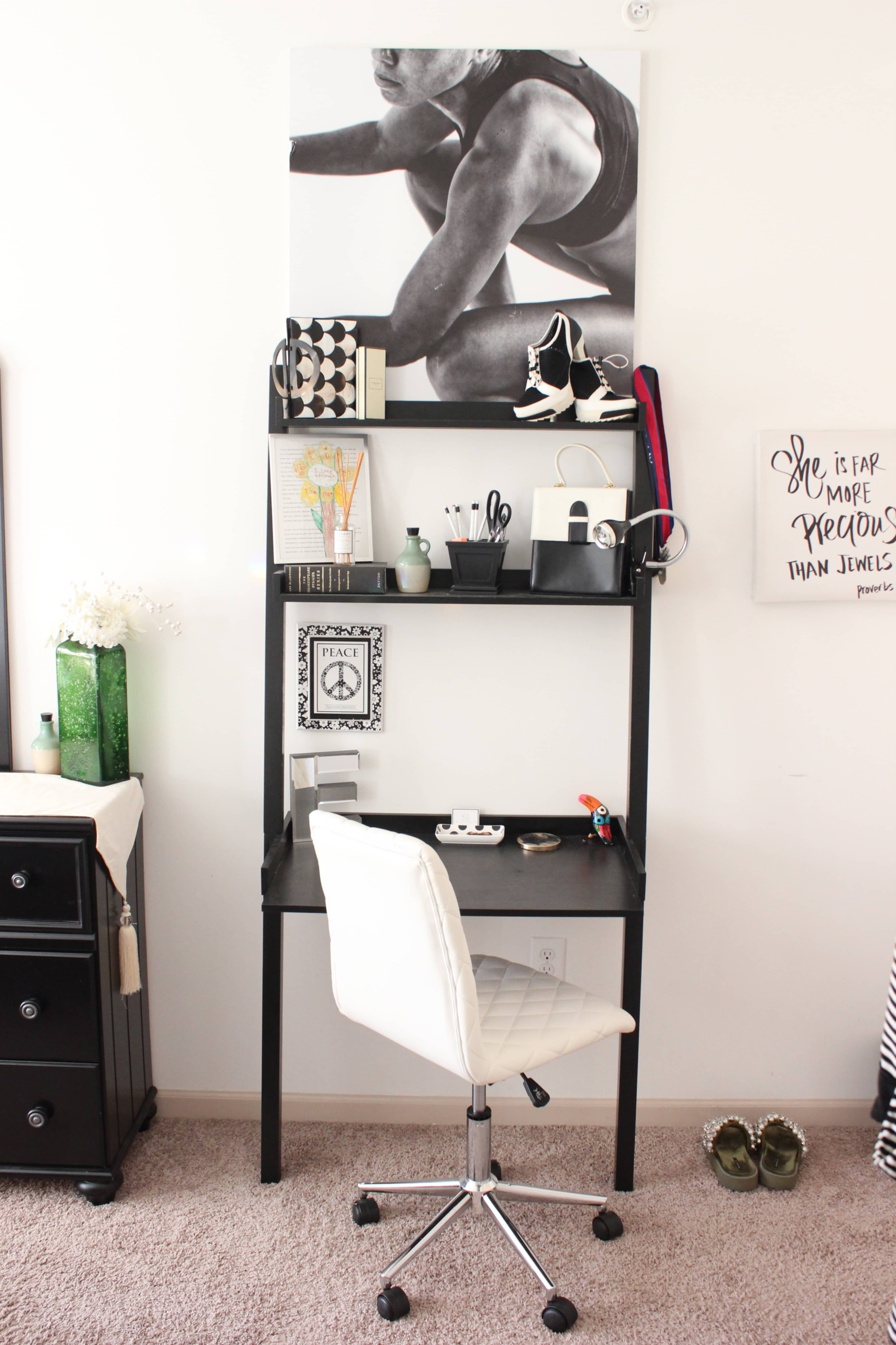 A minimalist desk setup features a black desk with a rolling chair, shelves displaying various decorative items and office supplies, and a large black-and-white poster on the wall above.