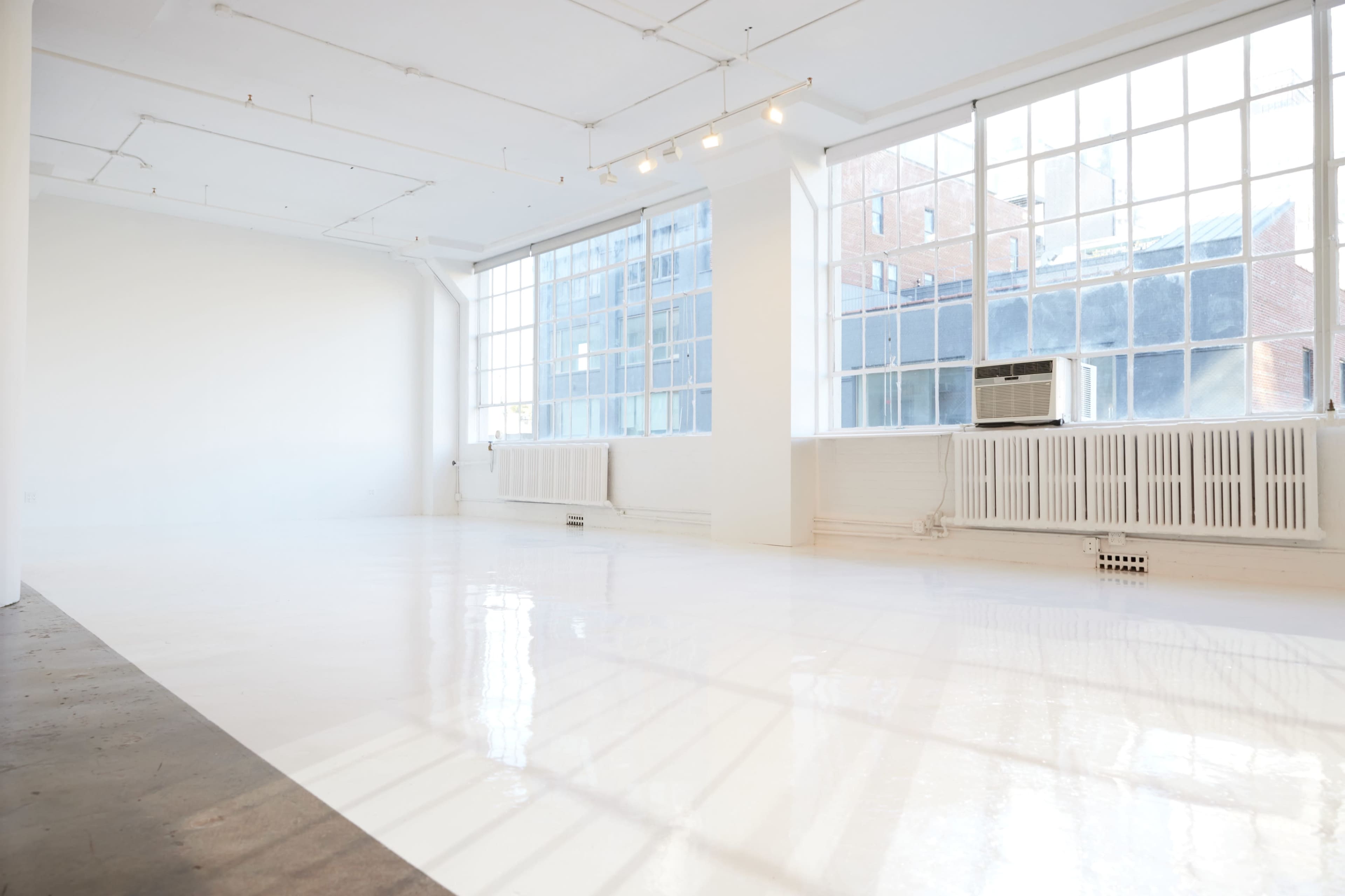 A spacious, empty room with large windows that provide natural light, featuring a polished white floor and industrial-style radiators along the walls.