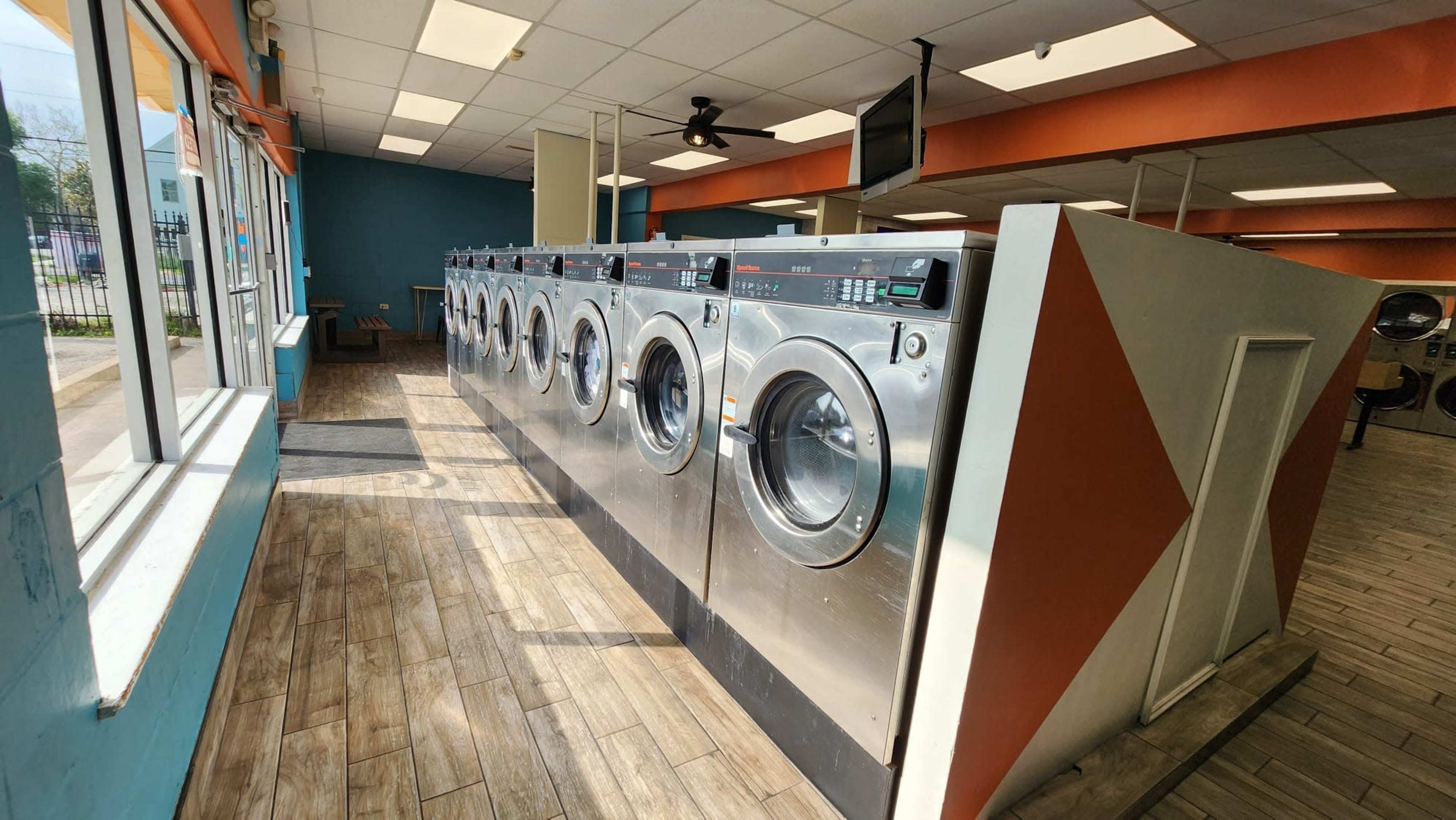 A row of commercial washing machines in a brightly lit laundromat with a blue and orange color scheme.