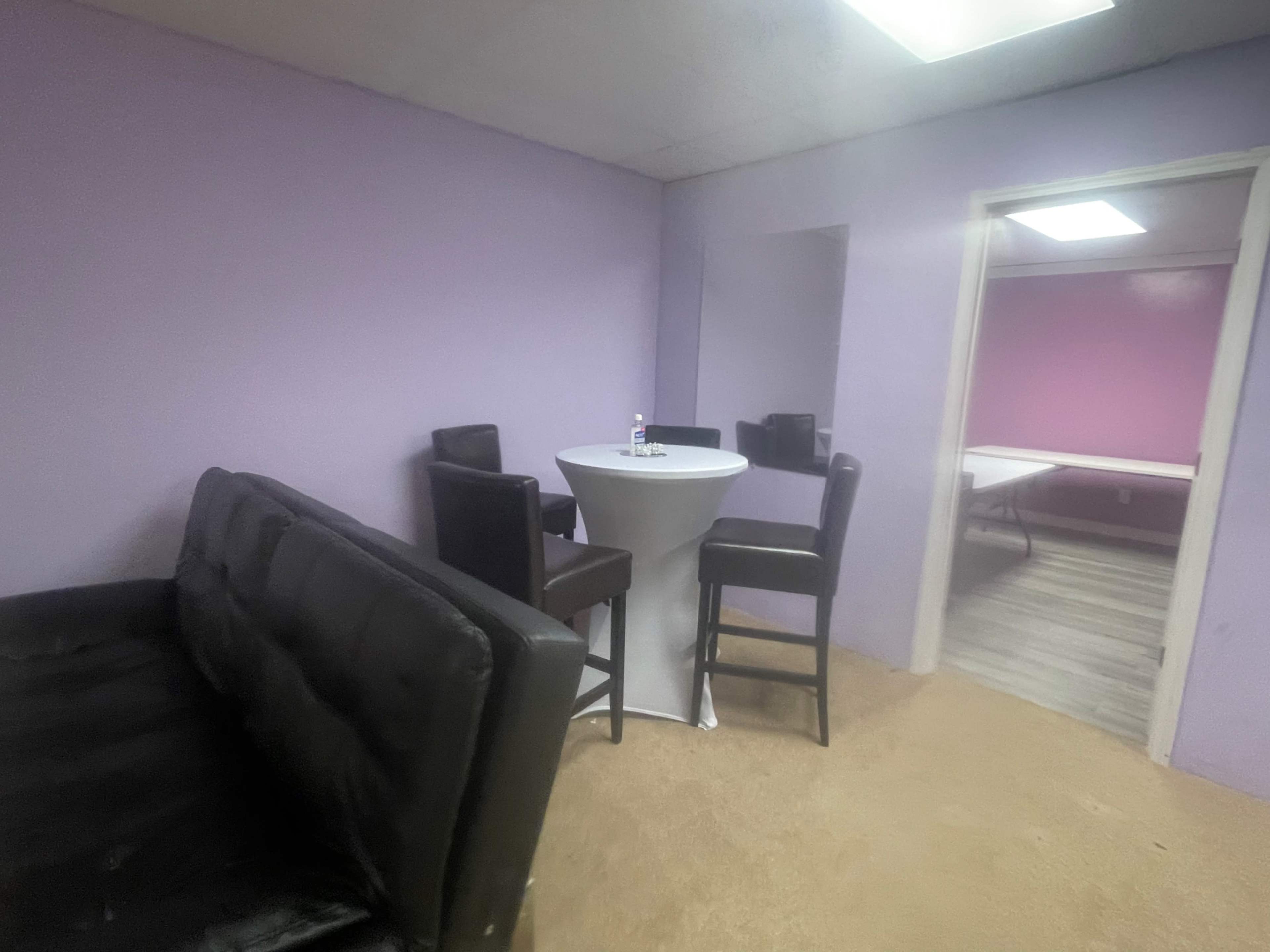 A small room with purple walls, featuring a round table surrounded by tall chairs, a black couch, and an adjoining room with a table visible.