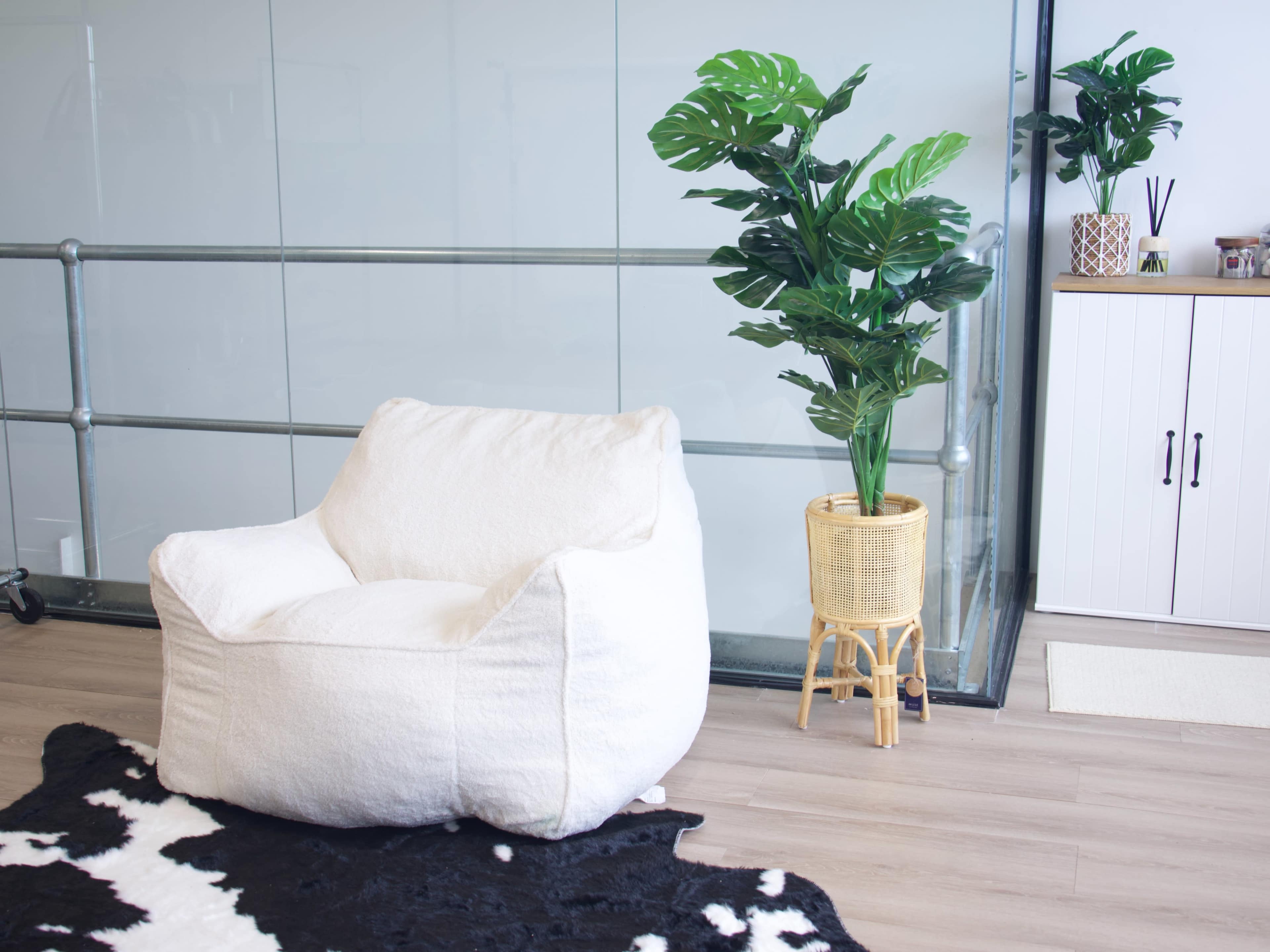 The image features a white bean bag chair next to a potted plant in a rattan planter, set against a glass wall and wooden floor.