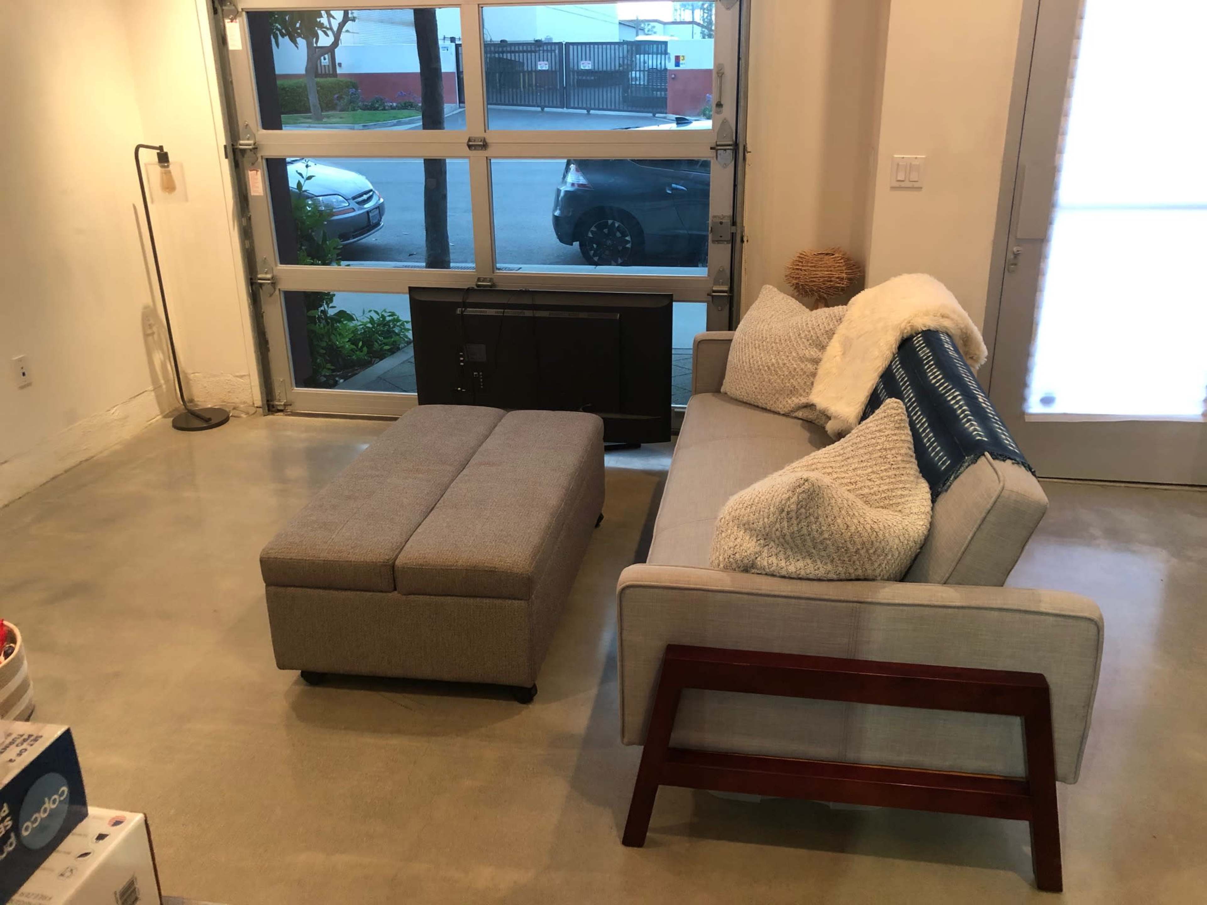 A minimalist living room with a gray sofa, an ottoman, and a large garage door opened to the outside.