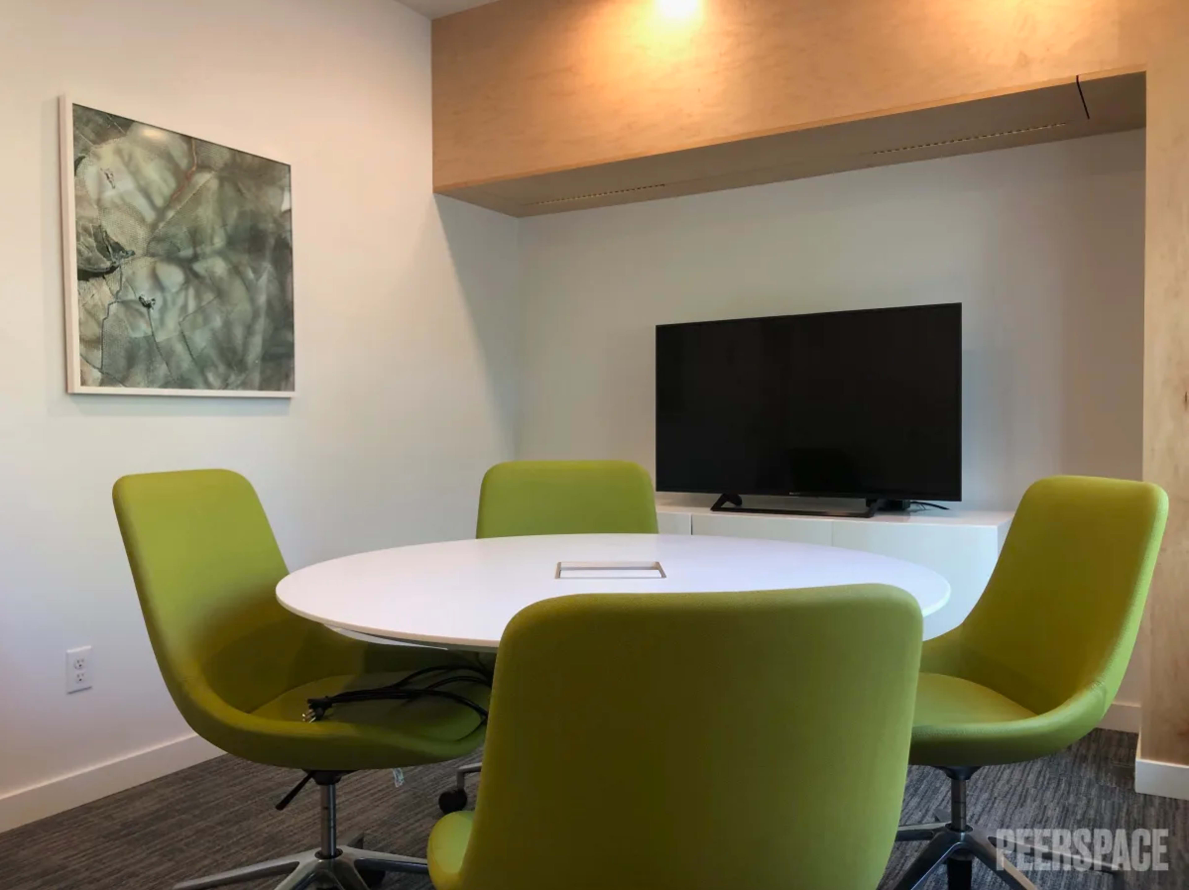 A round white table with four green chairs is set in a brightly lit conference room featuring a wall-mounted television and an abstract painting.