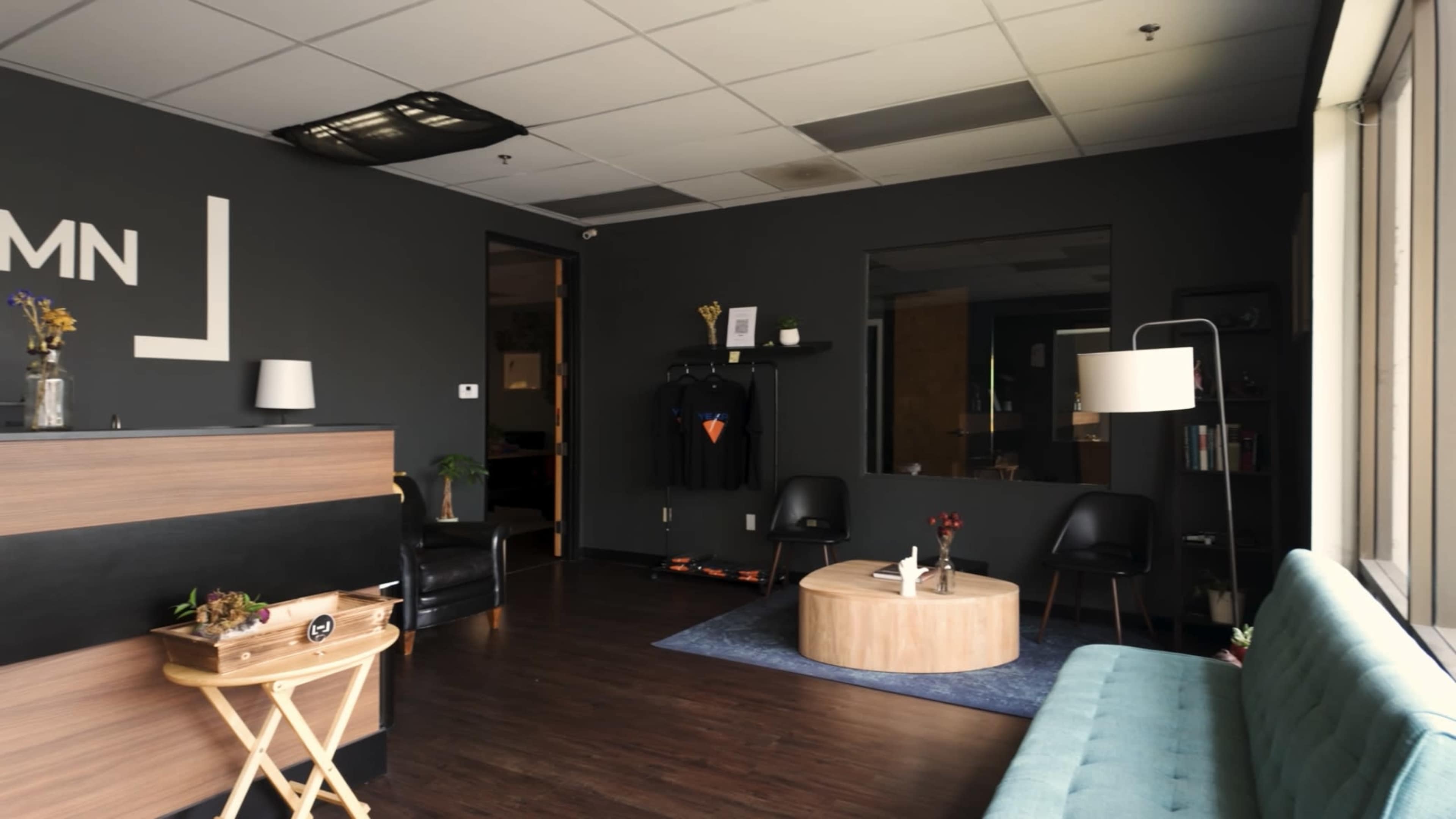 A modern office reception area with a wooden desk, a circular coffee table, a turquoise couch, and several black chairs against dark walls.